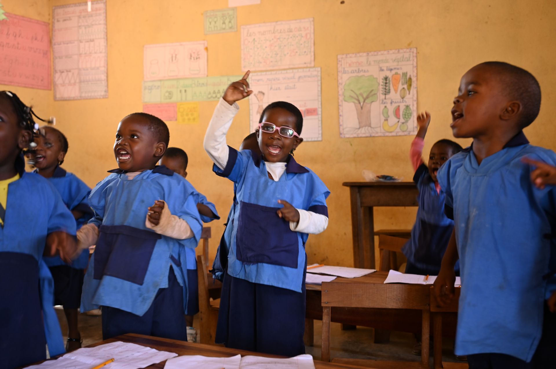 Children in a classroom standing up. One of the children is wearing a pair of pink eyeglasses