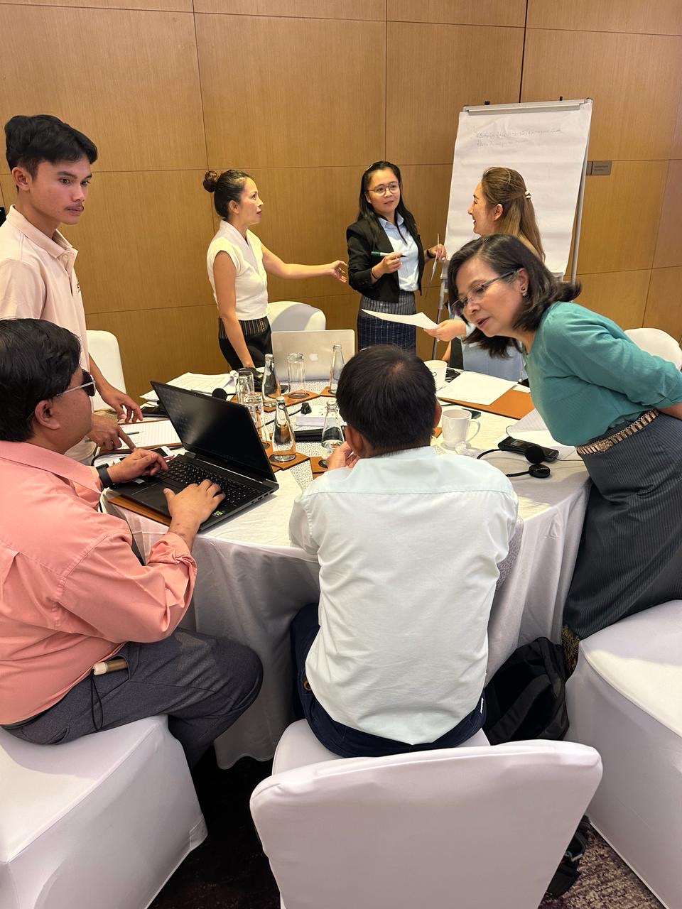 A group of people working with laptops around a table, some people are standing up and a white board in the background