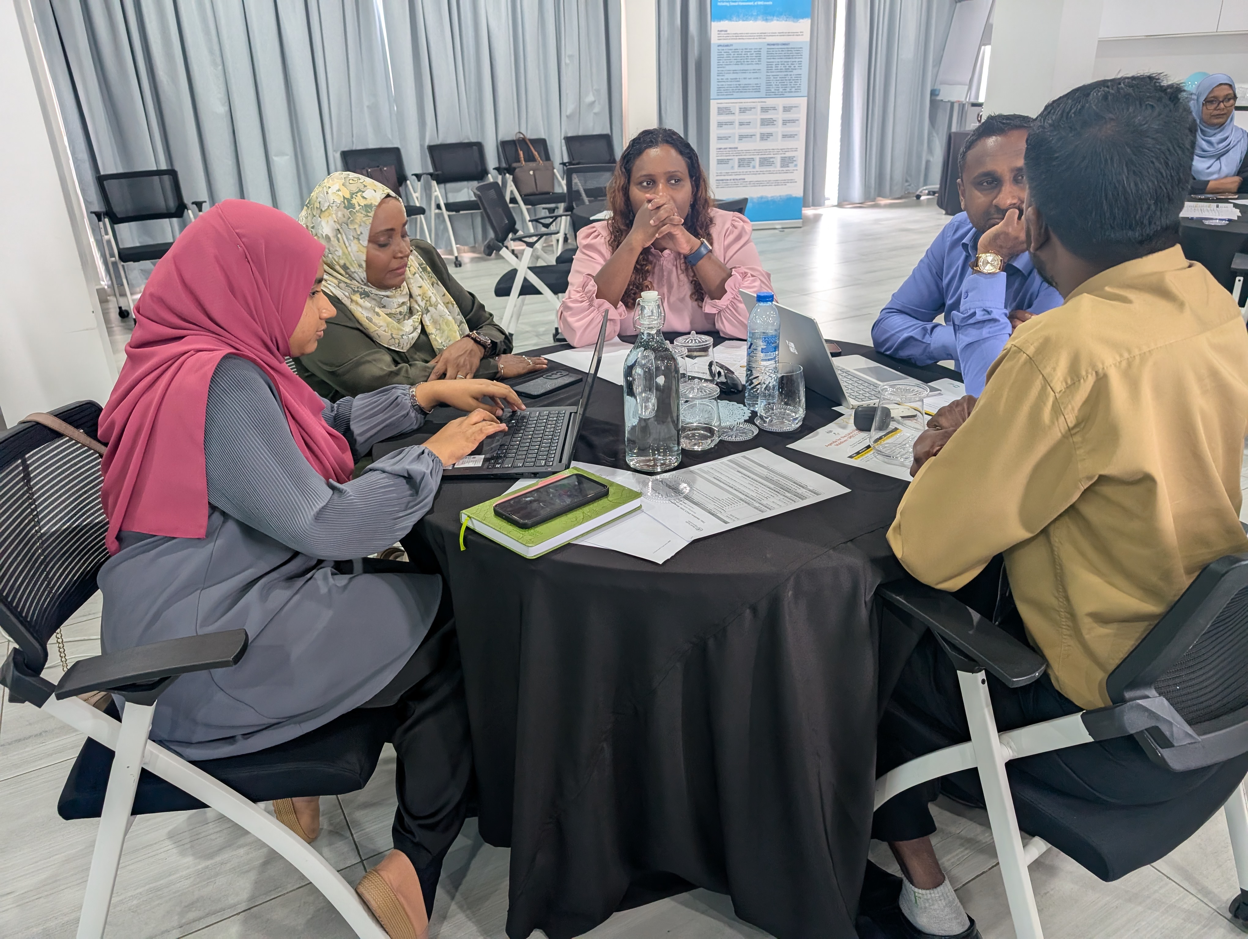 A group of people sitting around a table, laptops, mobile phones, bottles of water and glasses on the table.