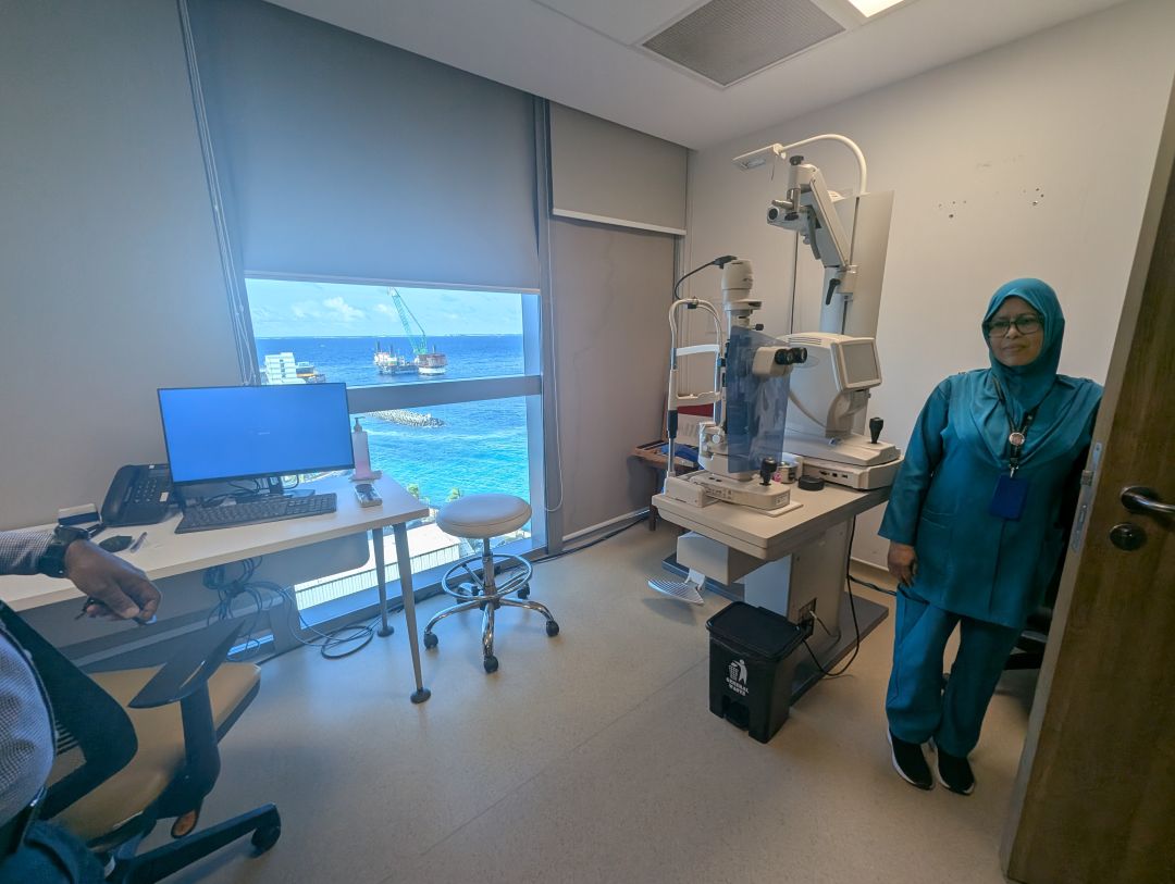 A woman in teal medical scrubs and headscarf stands next to eye examination equipment inside a clinic room. The room has a desk with a computer and a large window showing the ocean and construction barges outside.