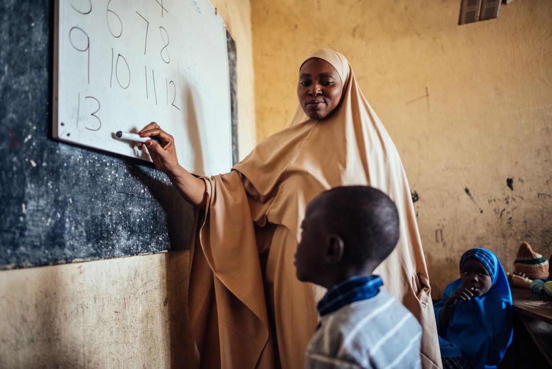 A teacher writing on a blackboard and a child reading out standing in front of the blackboard