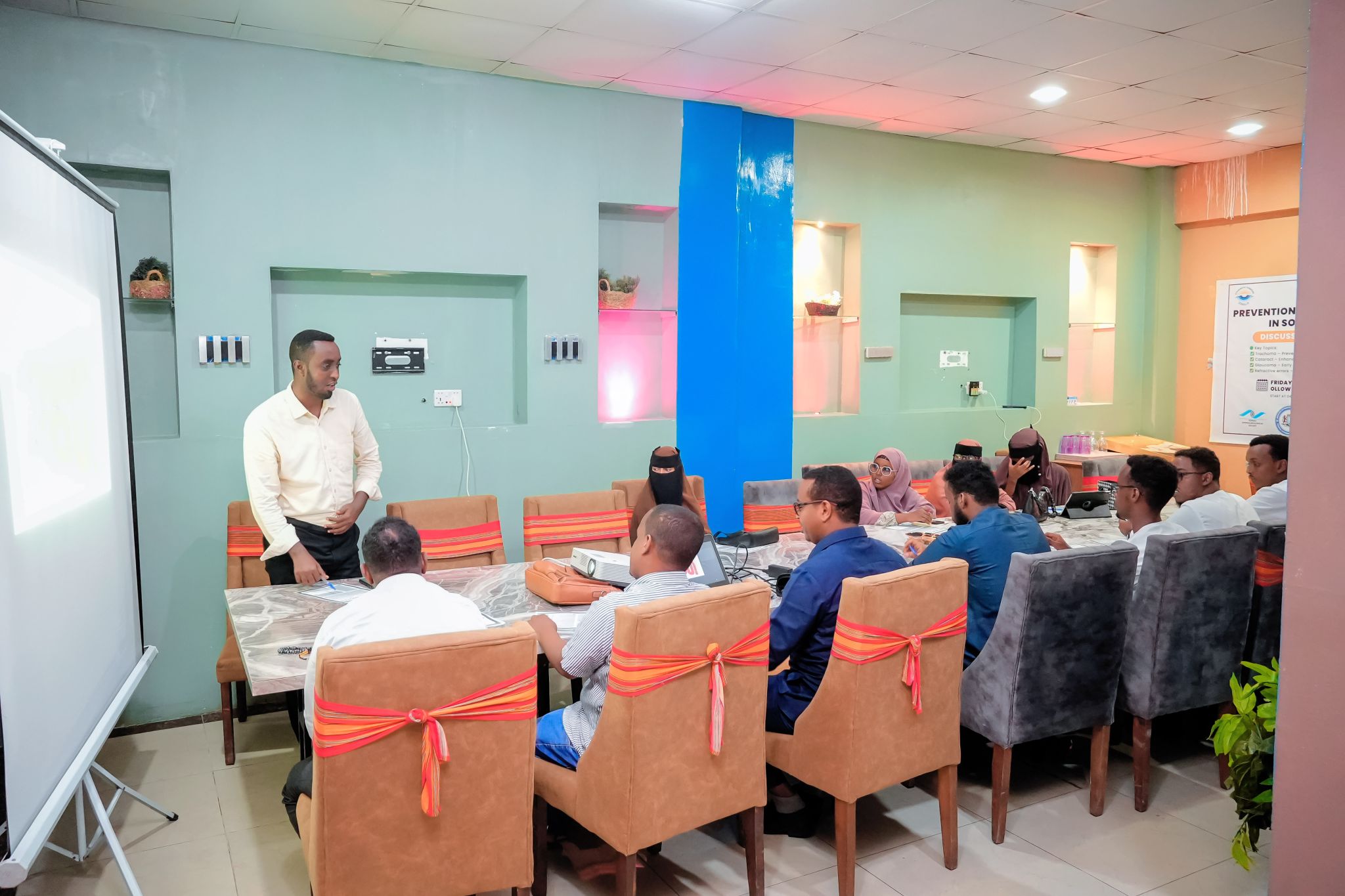 A meeting room with a long table with people sitting around it and a man standing up talking to them