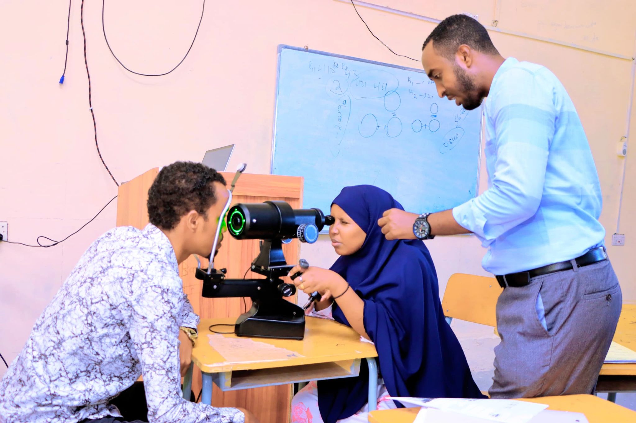 A man doing an eye examination to a woman and a second man standing up behind the woman