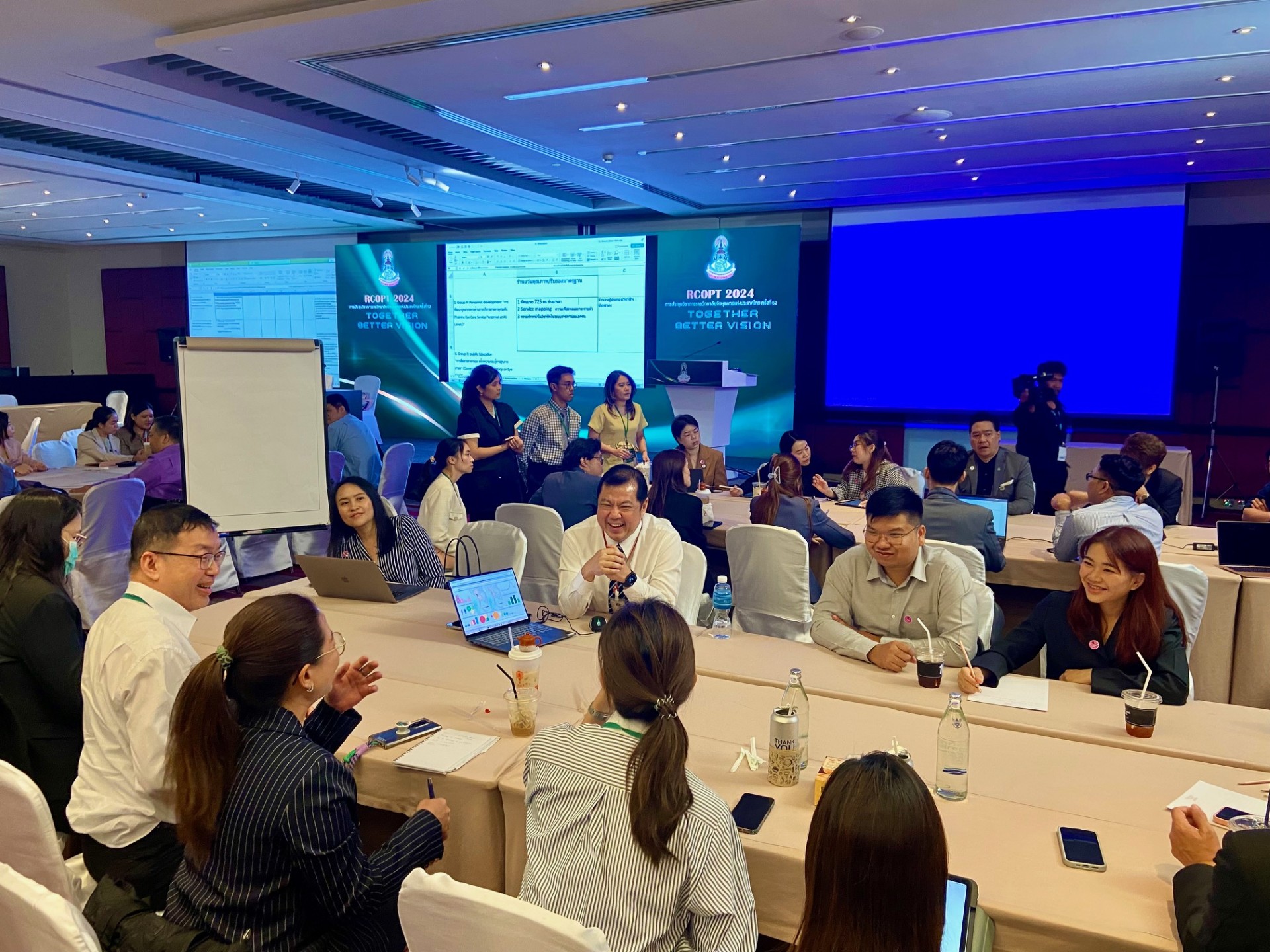 A group of people sitting around a table. Big screens at the background