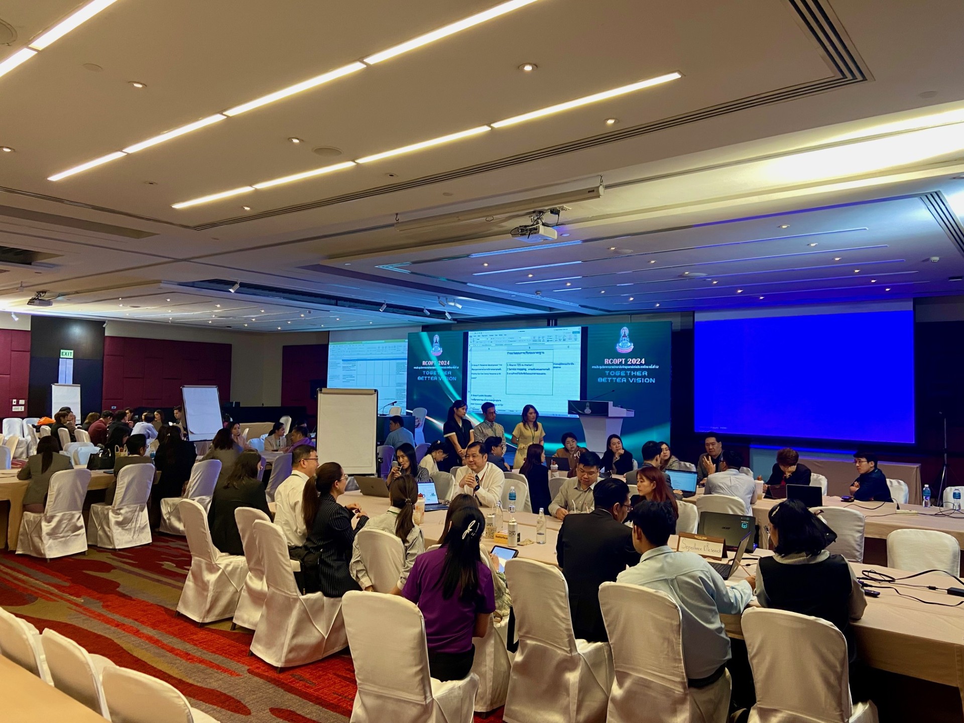 A meeting room with people sitting around a table and big screens at the background
