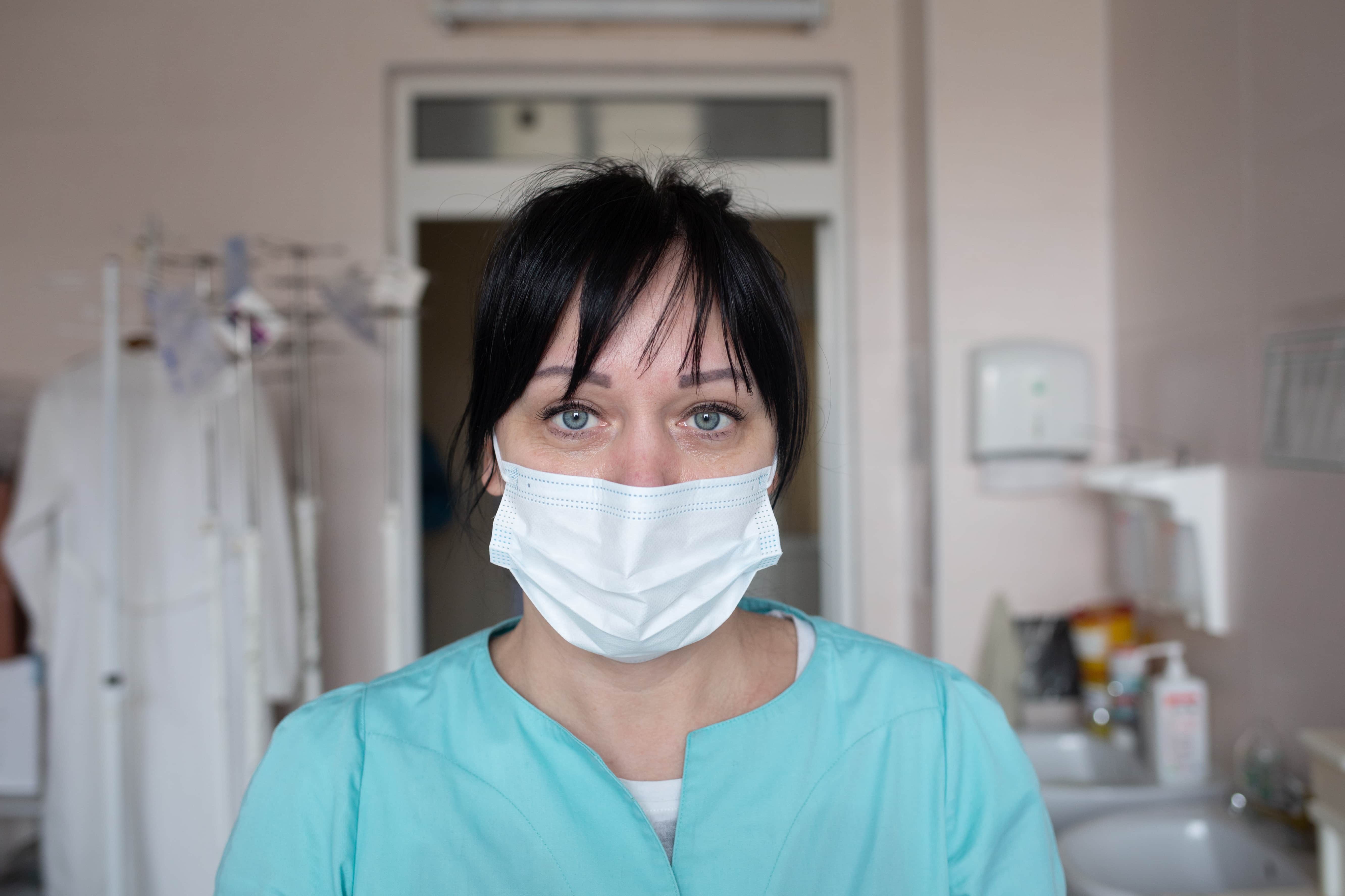 Portrait photo of a female nurse in a health care setting, wearing hospital attire and a face mask.