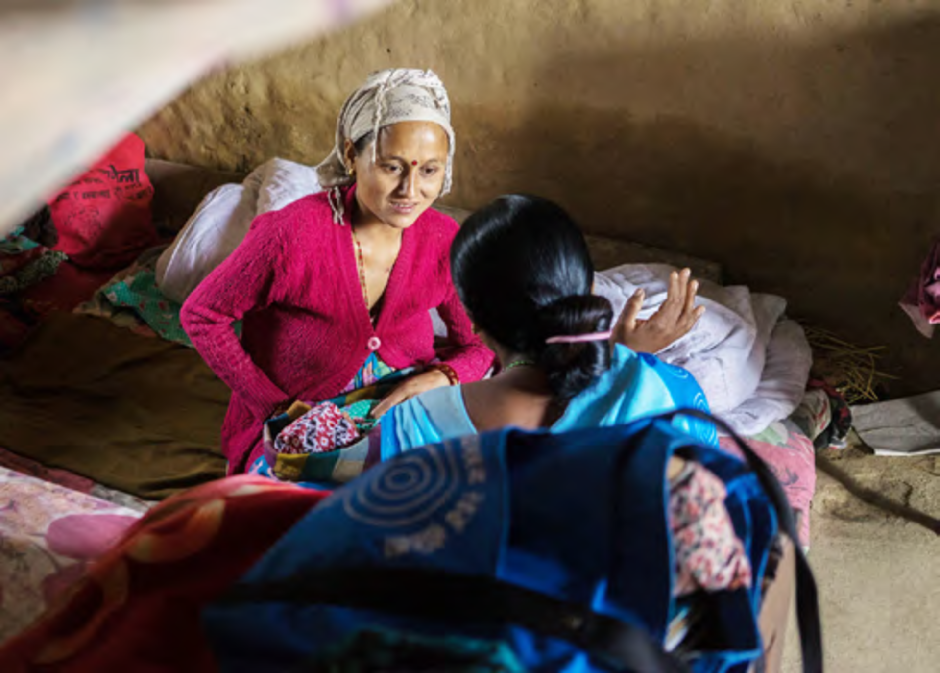 A midwife and a patient converse in a shared living space furnished with multiple beds and personal items.