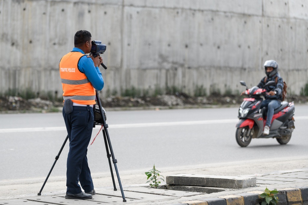 Traffic personnel in a reflective vest is standing at the side of a road holding a speedometer. A moped is seen driving on the road.