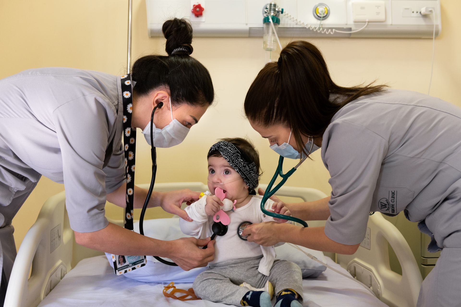 Two female doctors stand on each side of a baby, listening to her lungs with stetoscopes.