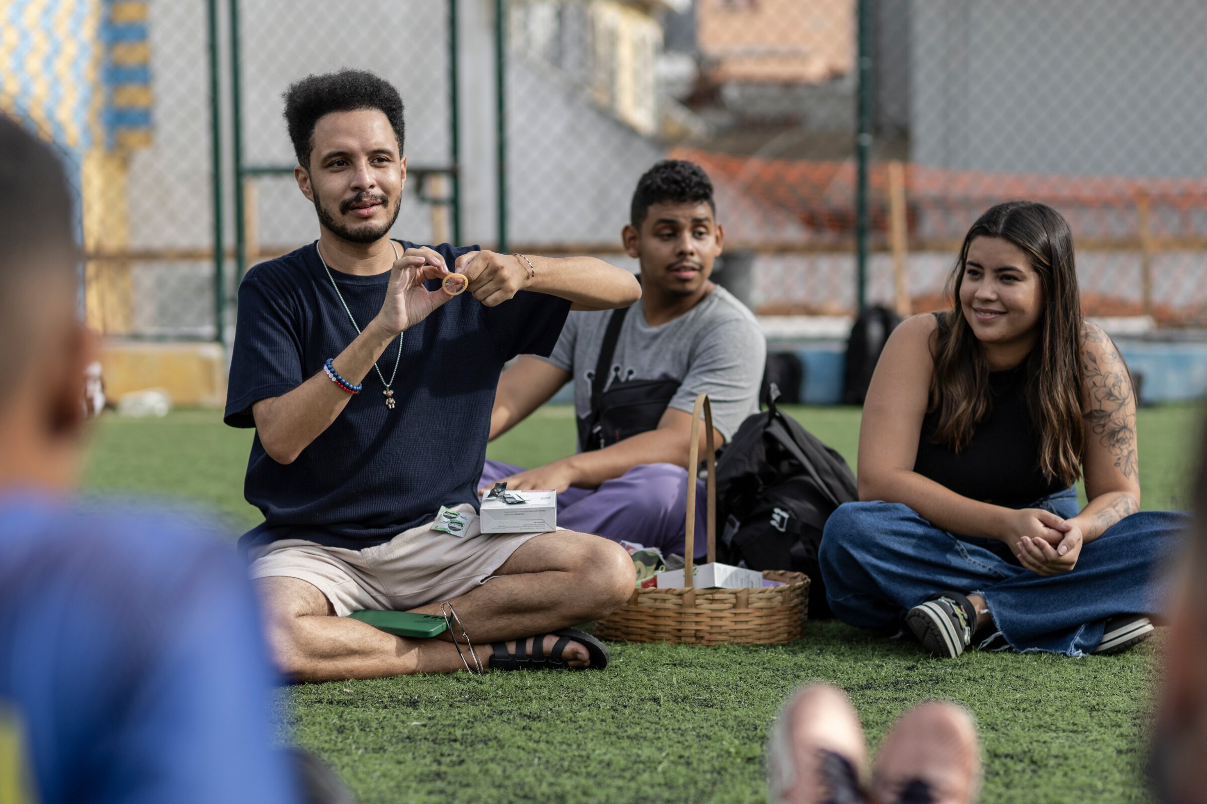 Three people sit outdoors on a sports court during an HIV and STI prevention activity, with one person demonstrating how to use a condom.