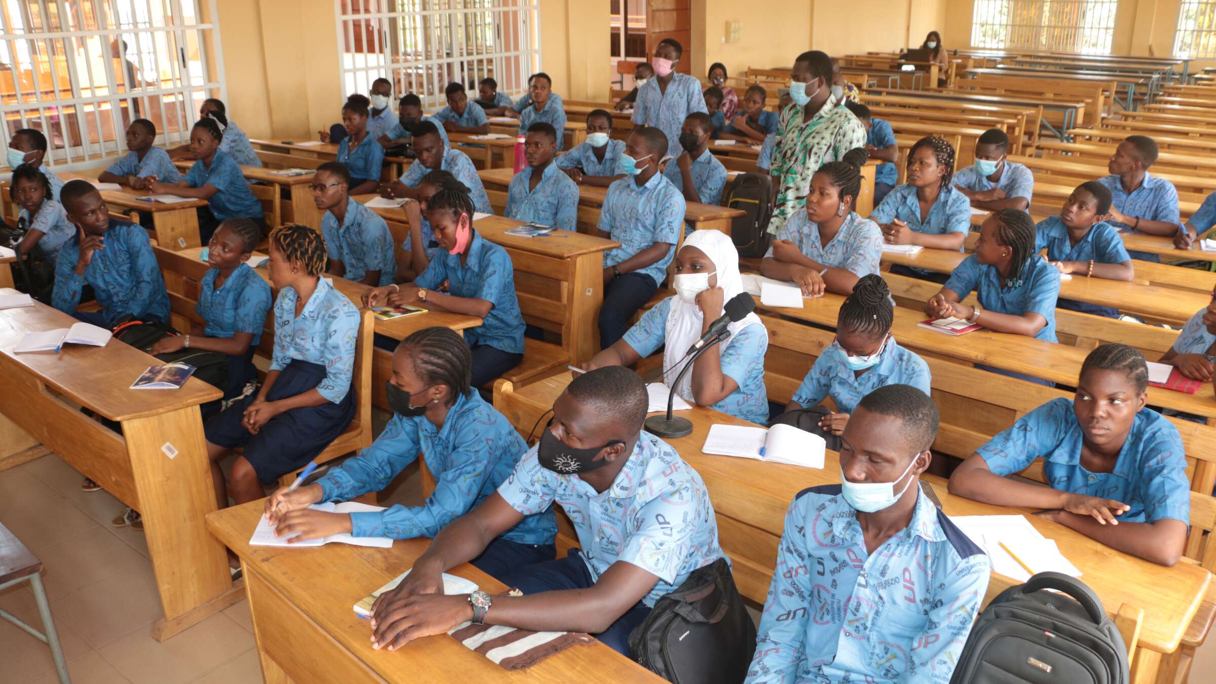 Students in blue uniforms sitting at a lecture, some wearing face masks whilst others are not.