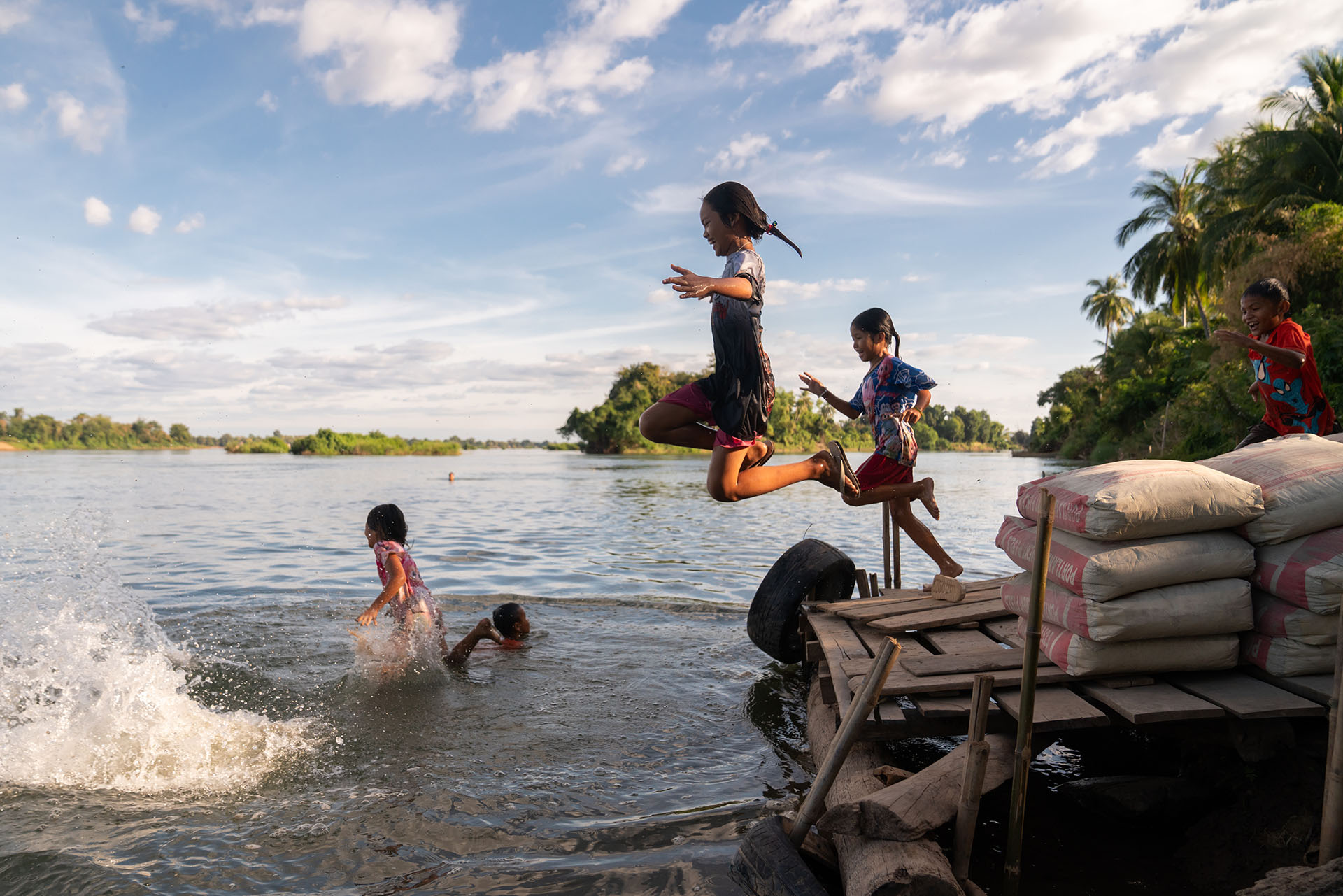 Children jump into the Mekong River from a wooden platform beside sandbags, with others swimming nearby.