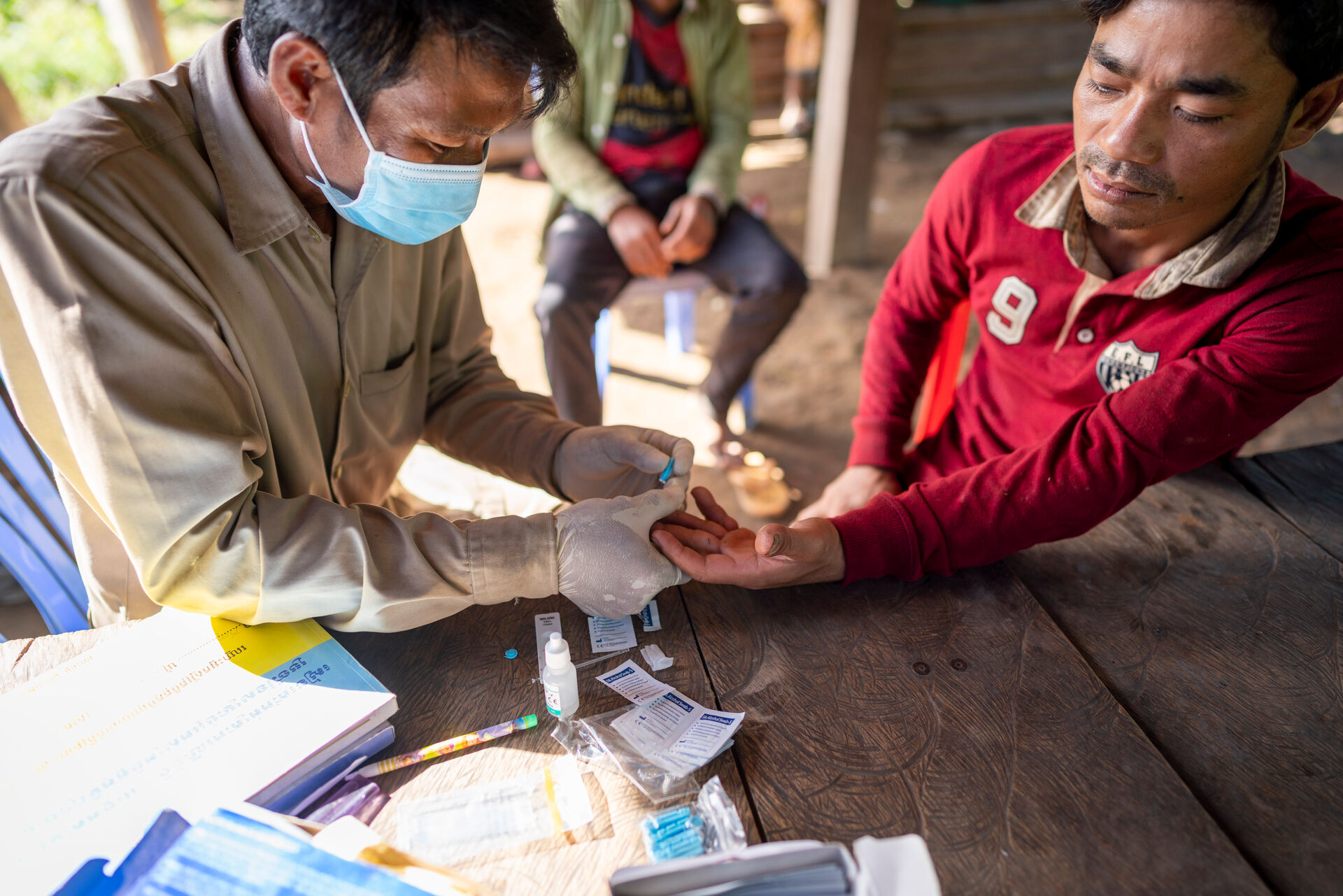 Masked person collects a finger-prick blood sample from another person during malaria testing at a wooden table.