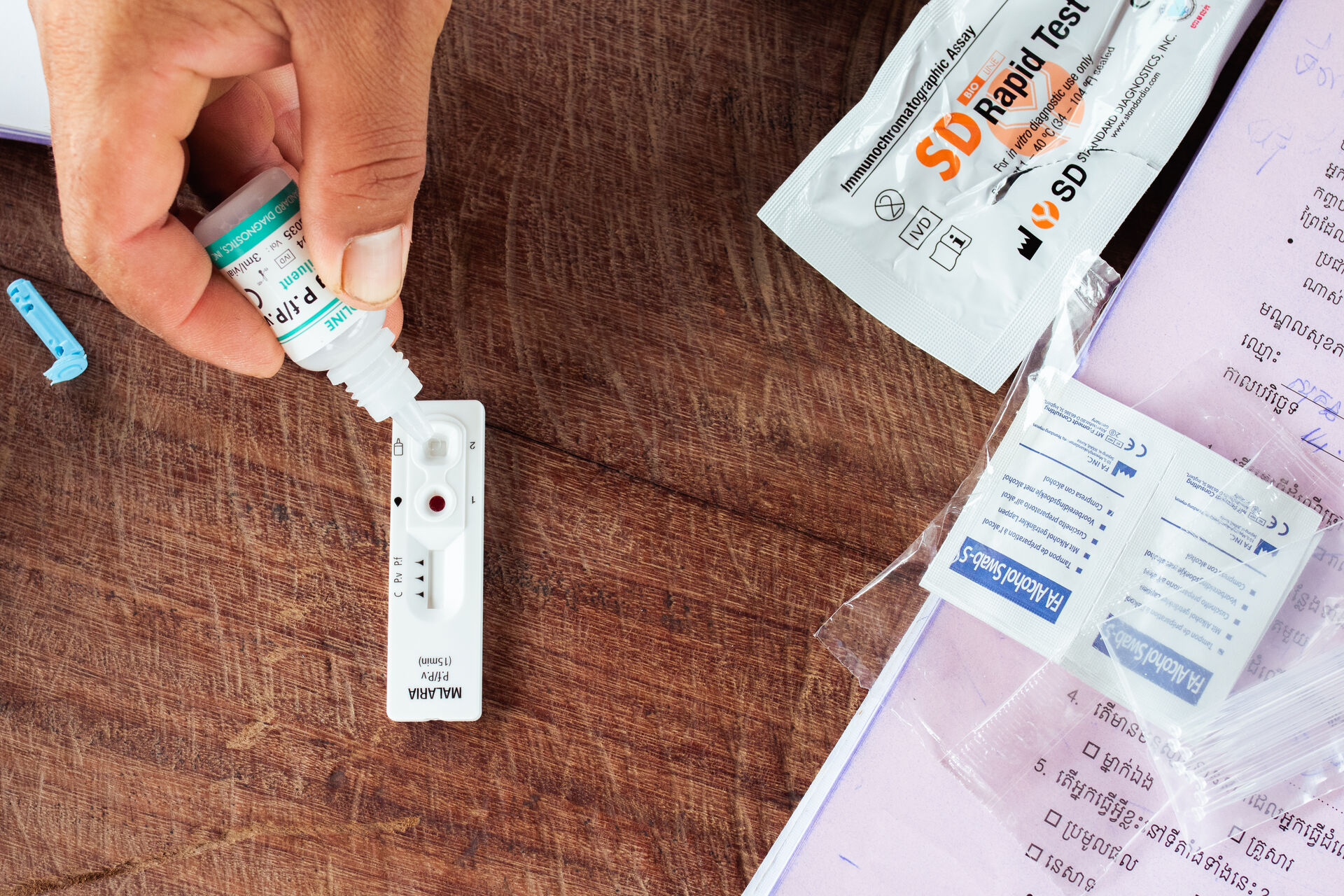Hand applying buffer drops to a malaria rapid diagnostic test cassette on a wooden table, with test kit materials nearby.