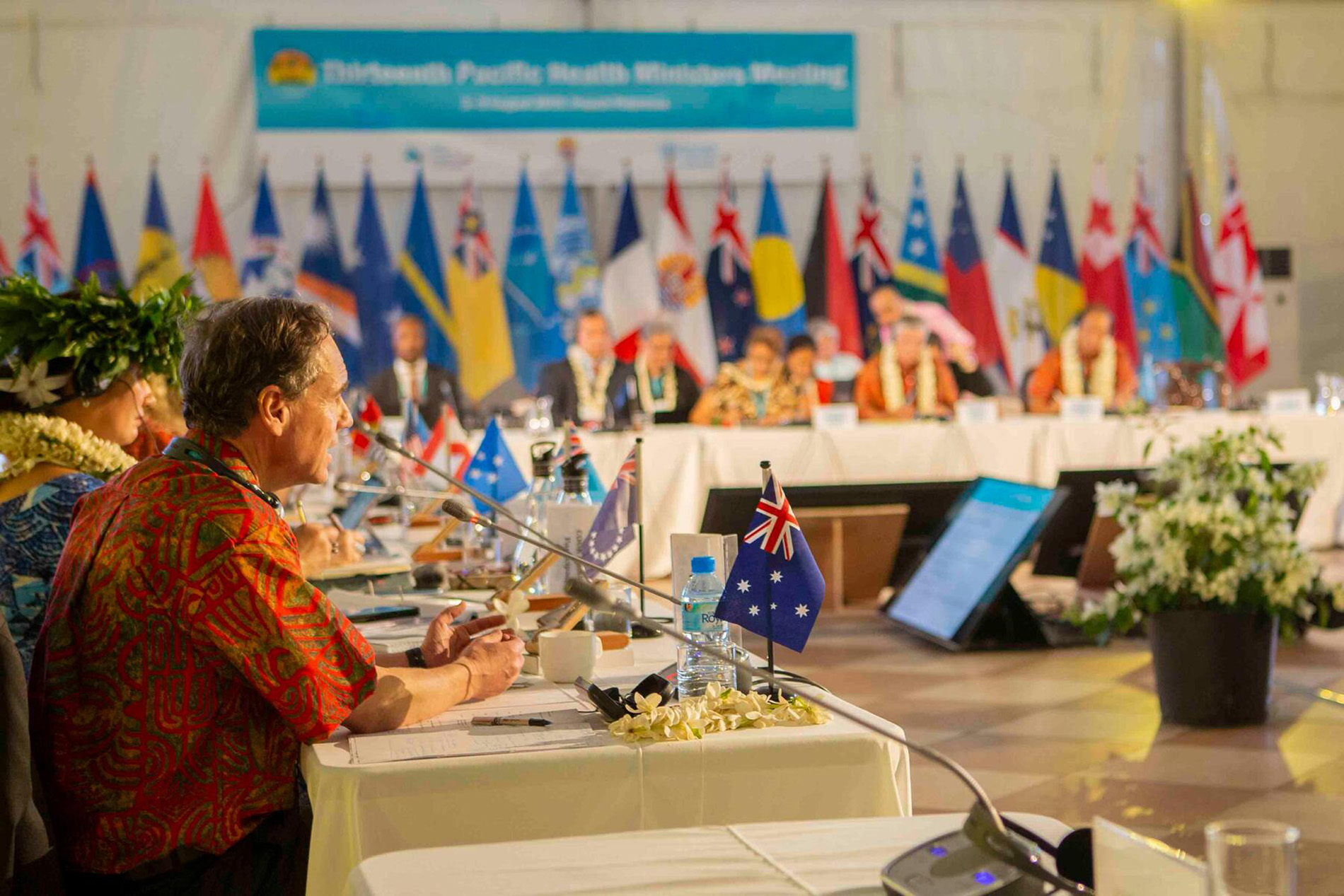 Delegates seated at a long table with microphones and country flags in a conference room, with a backdrop of national flags and a banner reading "Fifteenth Pacific Health Ministers Meeting."