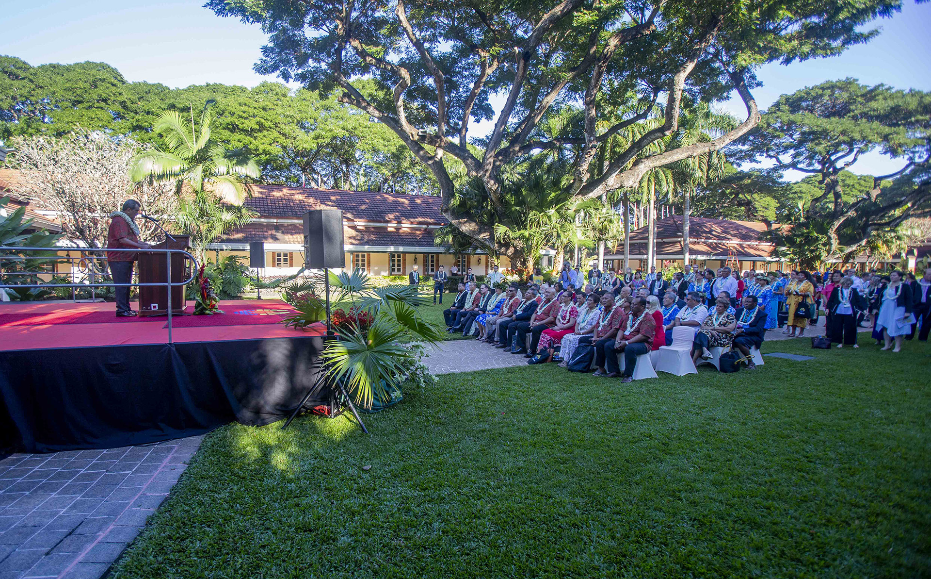 A speaker at a podium on a red-carpeted outdoor stage addresses an audience seated on white chairs, surrounded by greenery and trees, in front of a yellow building with a tiled roof.