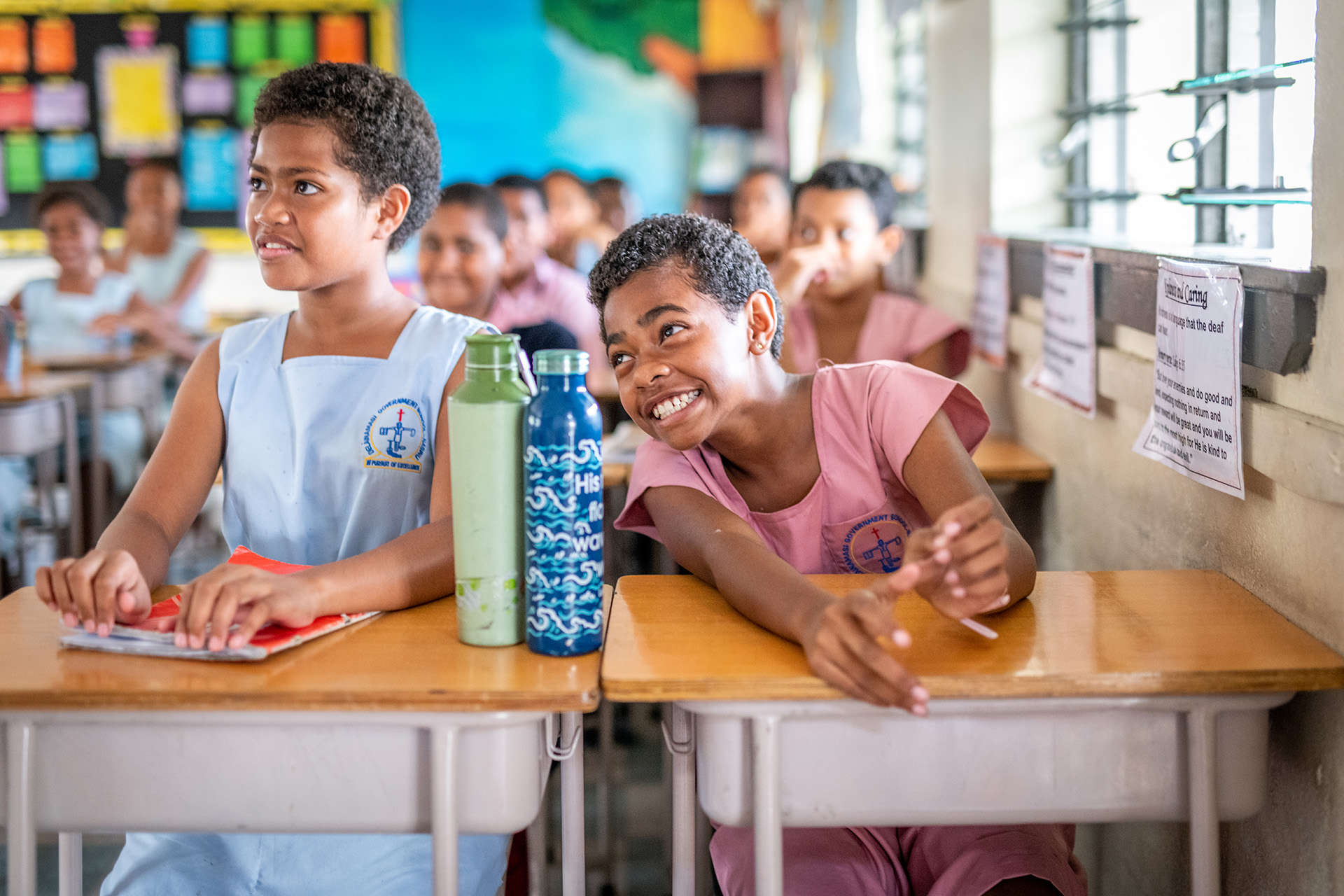 Classroom with students in blue and pink uniforms seated at desks, with water bottles on desks and colourful posters on the wall.