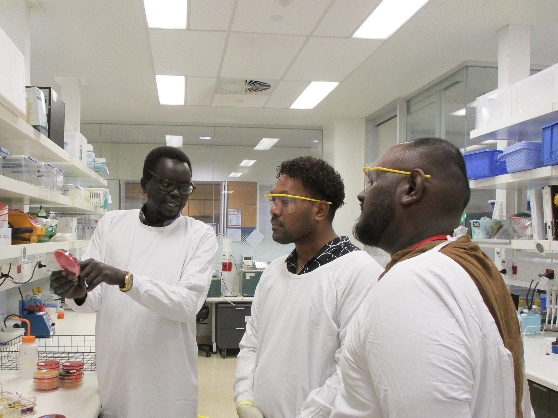 Three people in white lab coats stand in a lab as one shows a petri dish to the others. Shelves in the background hold scientific equipment and supplies.