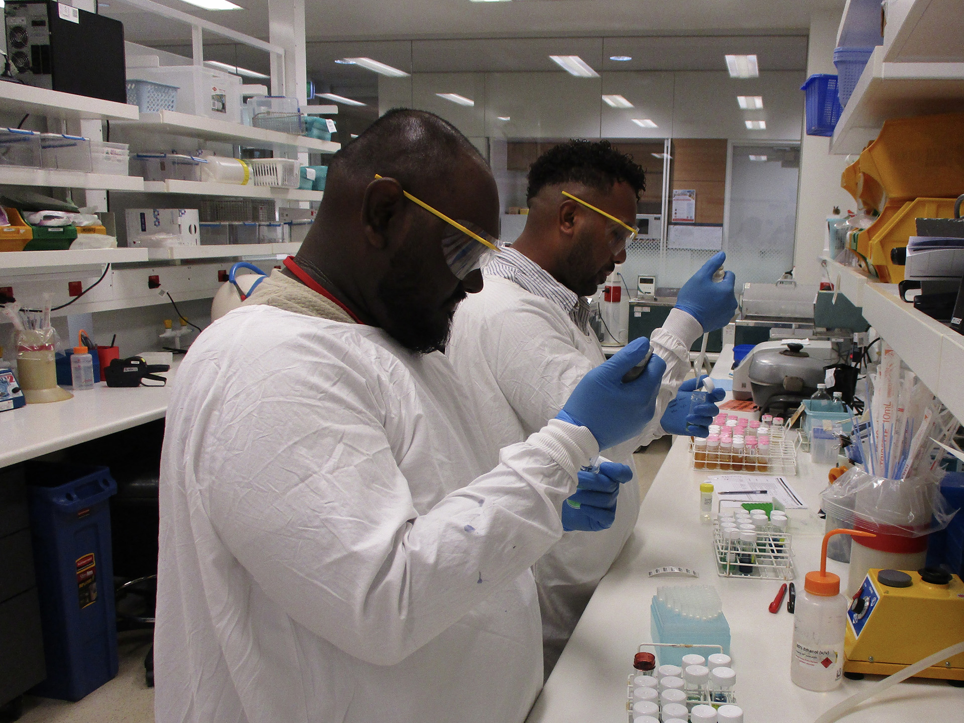 Two lab technicians in white coats and blue gloves use pipettes to transfer liquids into test tubes at a cluttered workbench surrounded by scientific instruments and supply shelves.