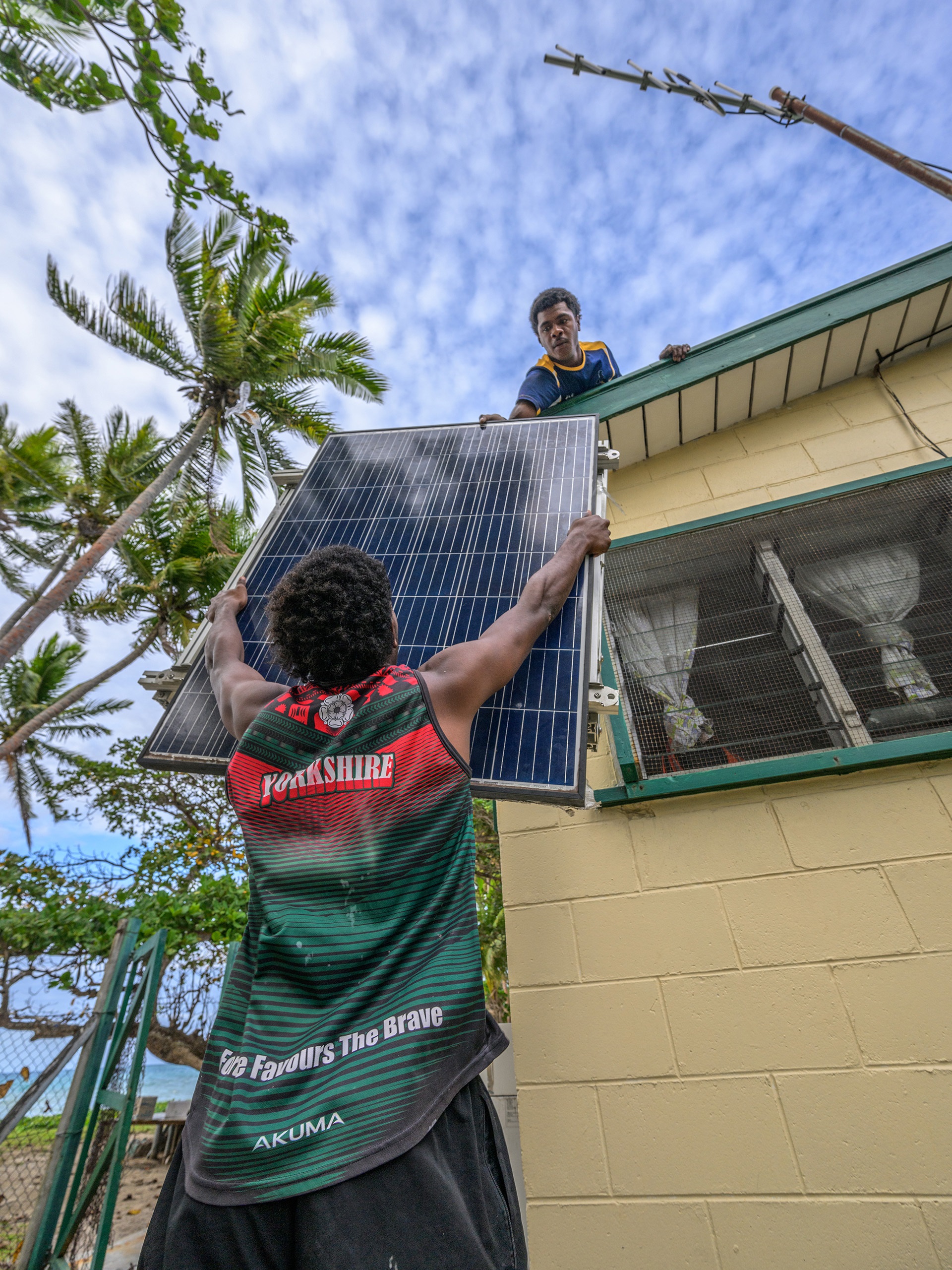 Coconut halves drying on a corrugated roof near thatched huts, palm trees, and hanging laundry in a tropical village.