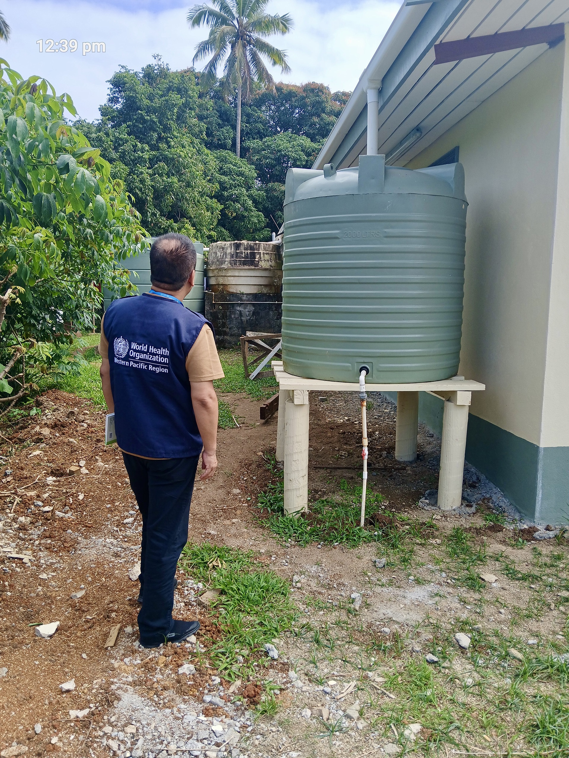 Coconut halves drying on a corrugated roof near thatched huts, palm trees, and hanging laundry in a tropical village.