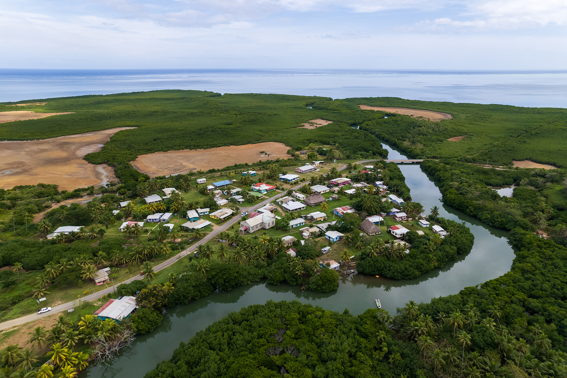 Aerial view of Lomawai, a coastal village with colourful rooftops, winding roads, lush greenery, and waterways, bordered by forest and fields, with the ocean in the background.