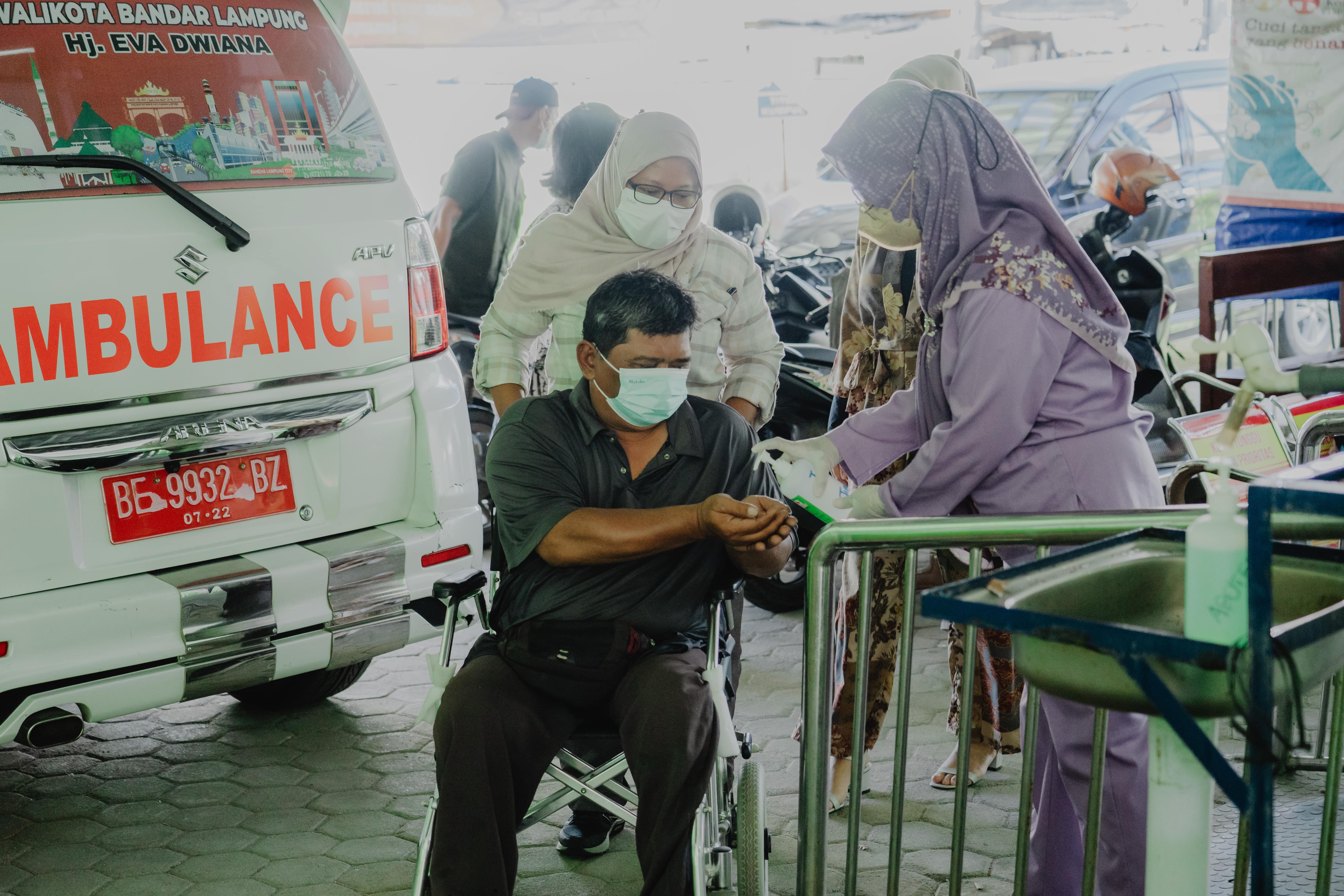 A man in a wheelchair has his hands sanitized by a health worker in front of an ambulance.