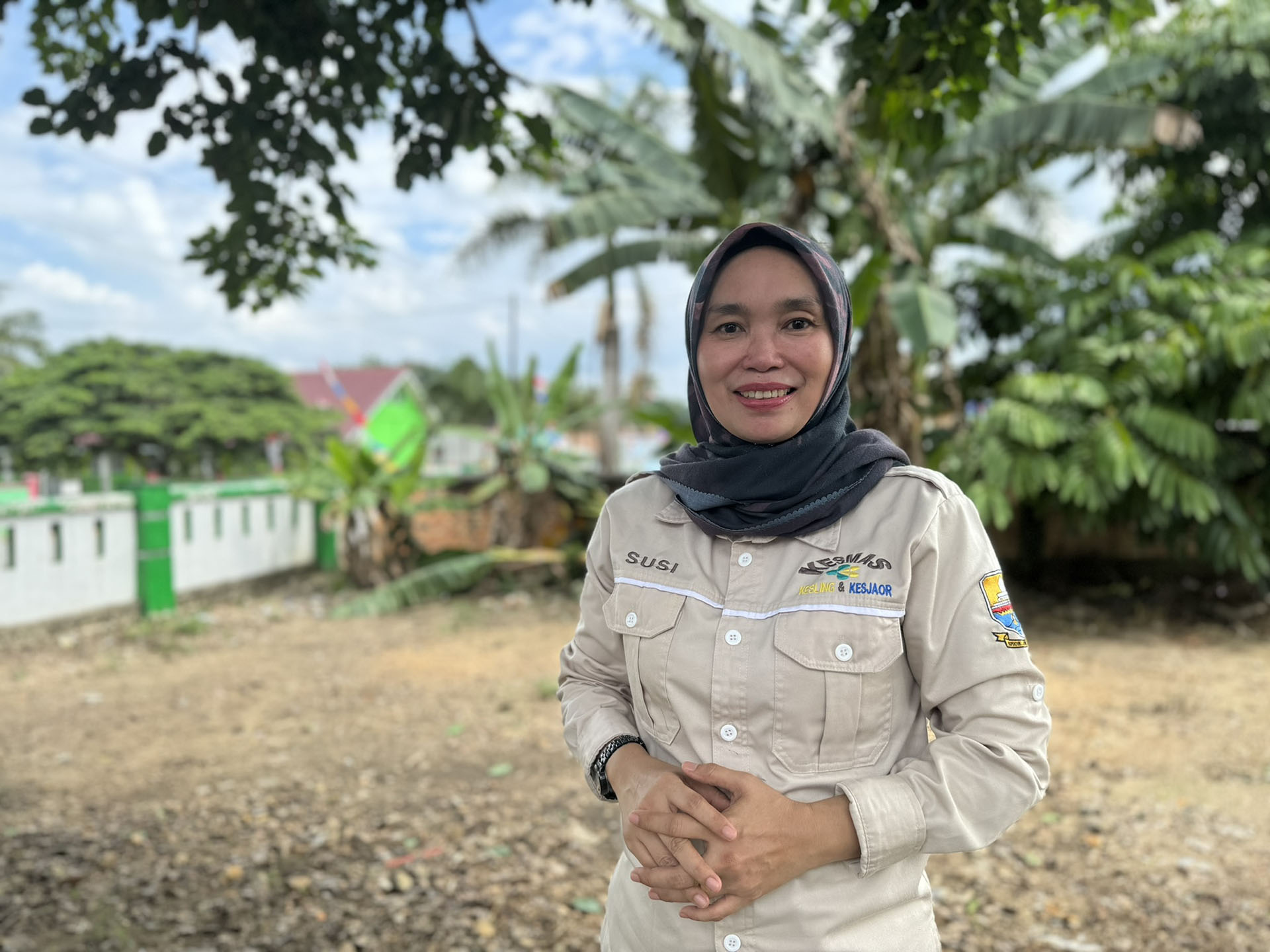 Person in beige uniform with name tag standing outdoors near trees and greenery.