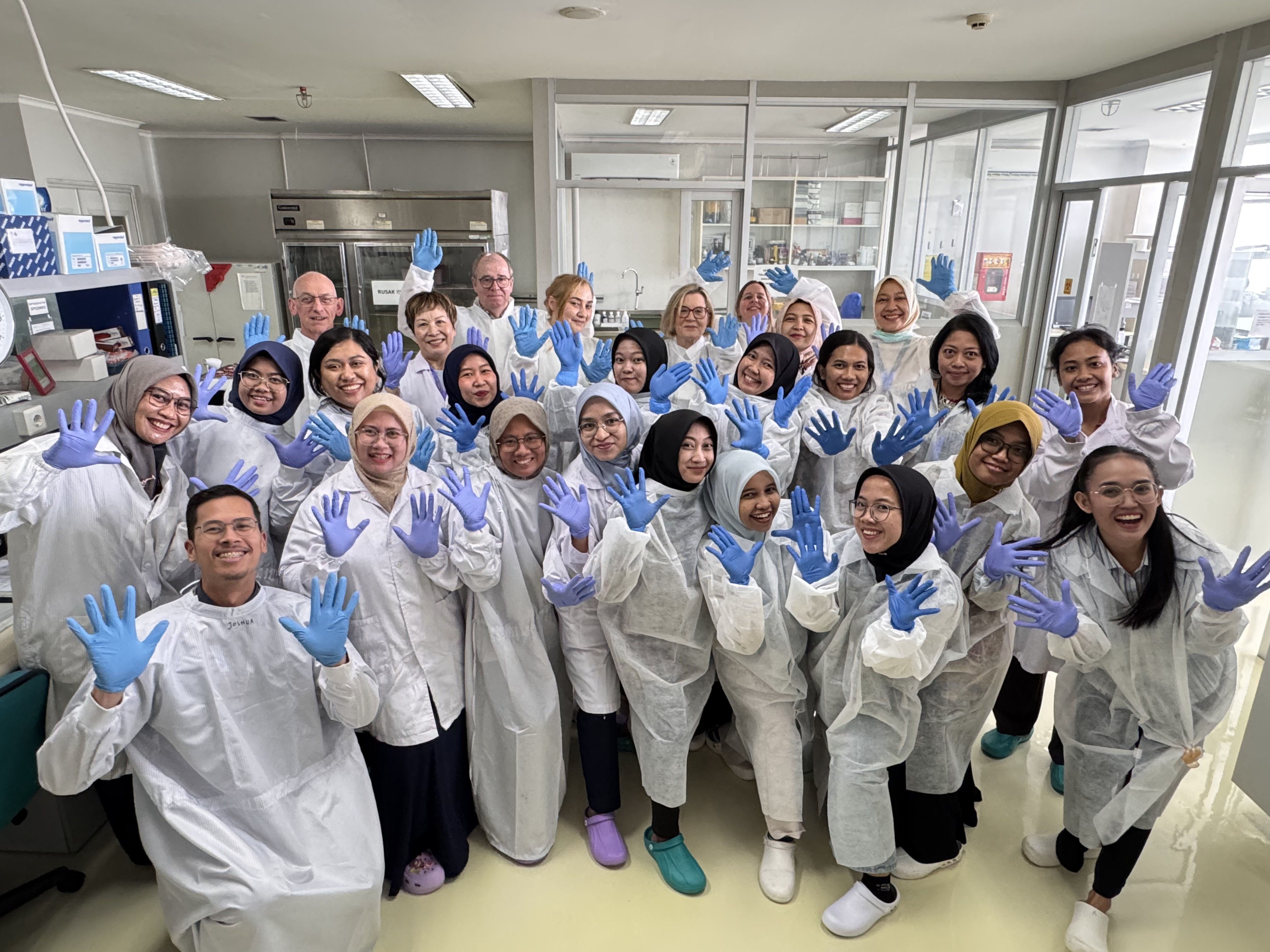 A group of laboratory training participants wearing protective gowns and blue gloves posing together in a laboratory room.