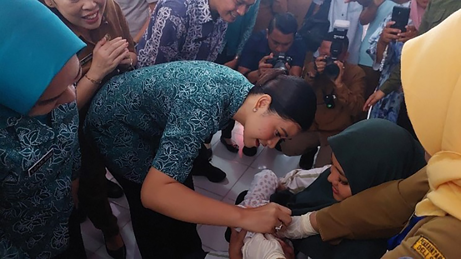Sekelompok orang dengan pakaian beragam berkumpul di sekitar wanita berkerudung hijau tua yang duduk sambil menggendong bayi. Seseorang berseragam toska membungkuk berinteraksi dengan bayi, sementara yang lain berdiri di dekatnya.