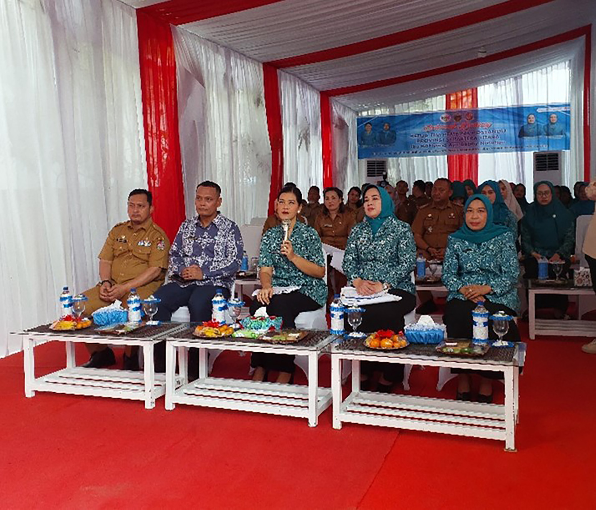 A group of people is seated, the front row consists of five individuals—three men and two women—sitting on white chairs behind small white tables with various snacks and drinks.