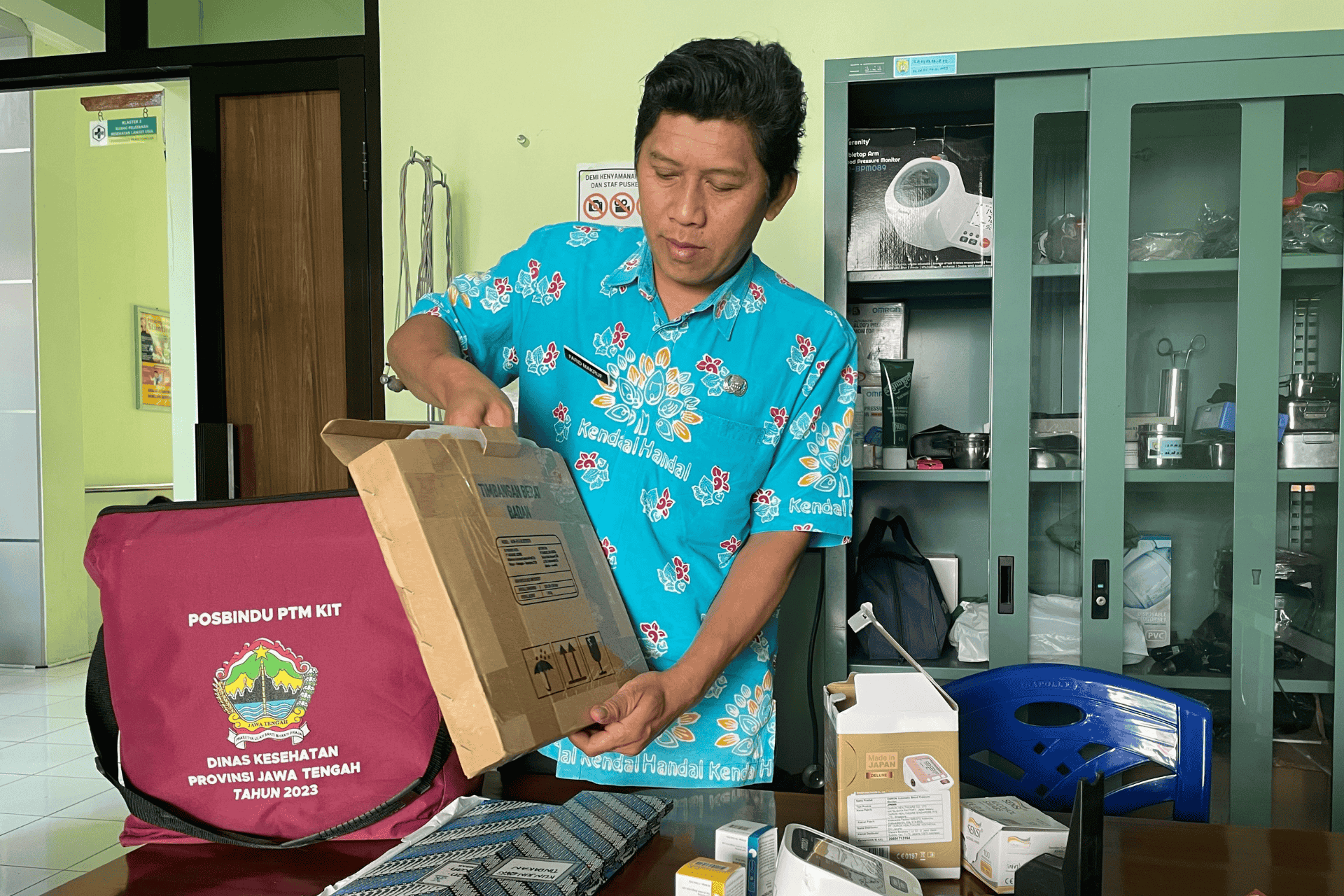 Orang membuka kotak “Timbangan Badan” di samping tas marun POSBINDU PTM KIT dan perlengkapan medis.