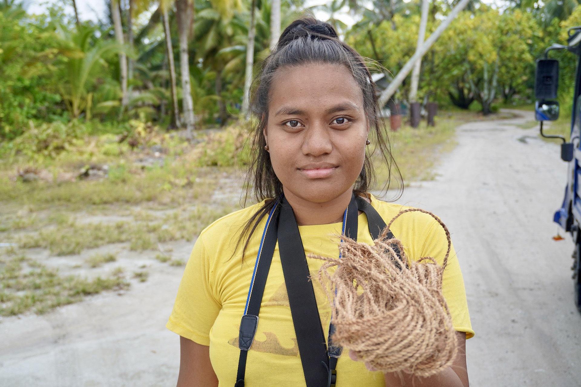 Coconut halves drying on a corrugated roof near thatched huts, palm trees, and hanging laundry in a tropical village.