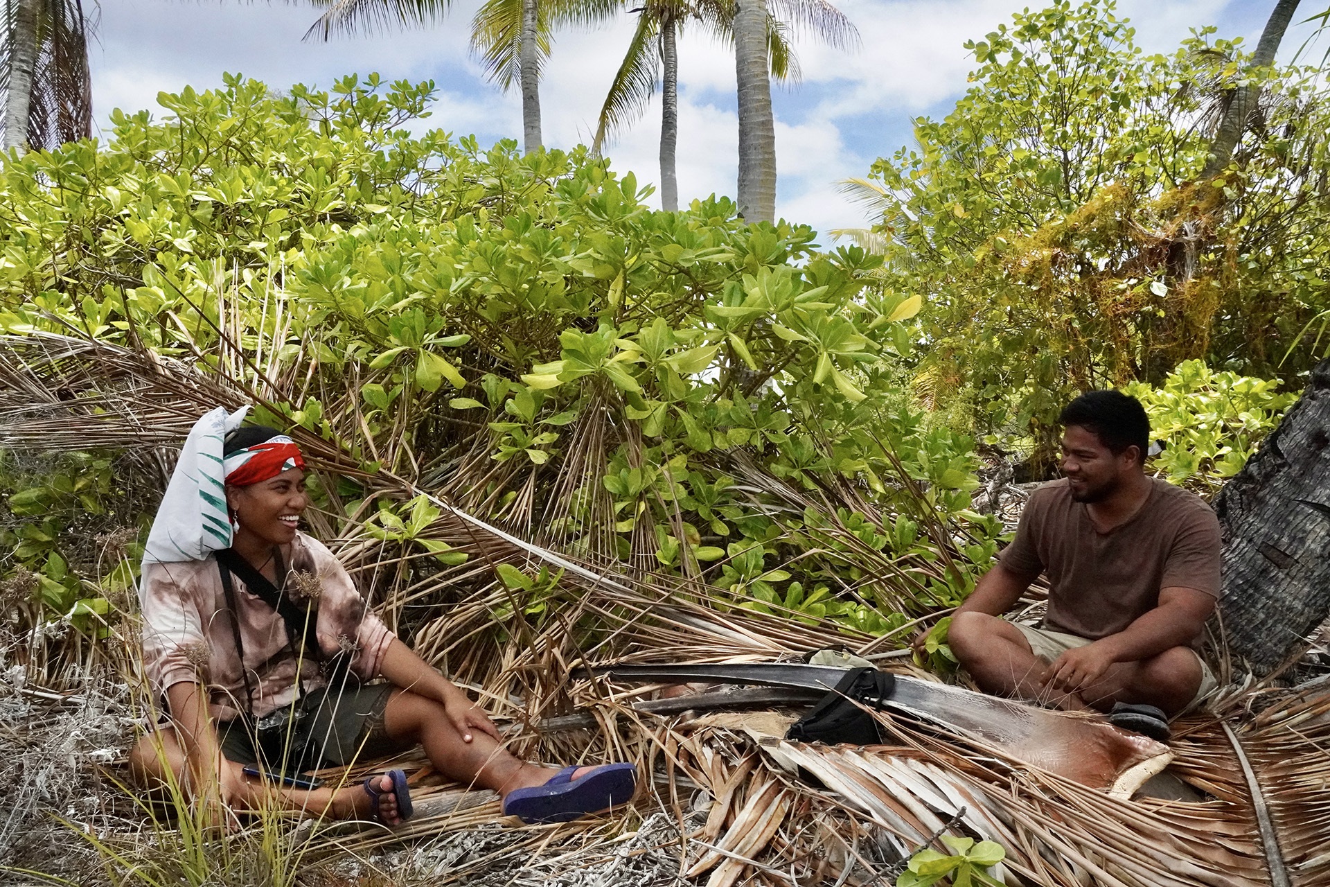 Two people sitting on dried palm fronds in a tropical setting with green shrubs and palm trees.