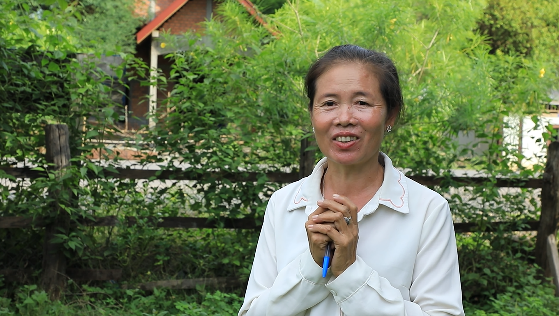 Person in a white collared shirt standing outdoors, holding a blue pen with both hands, in front of a wooden fence and lush green foliage with a house partially visible in the background.