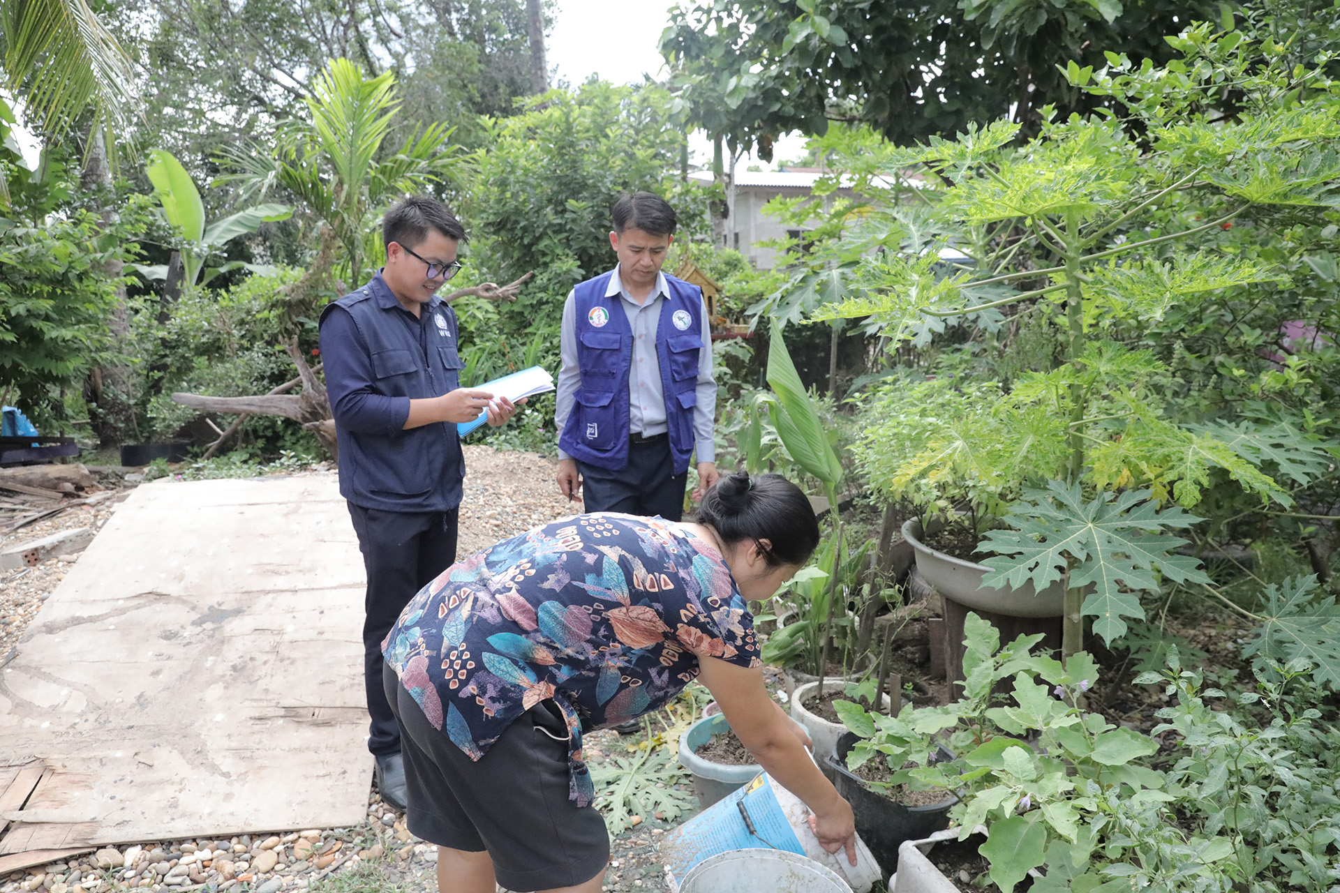 Two people in blue uniforms holding documents observe while another person tends to potted plants in a lush outdoor garden area with trees and greenery.