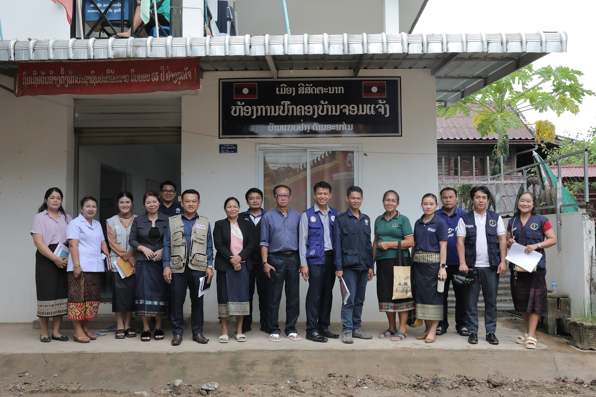 Group of people standing in front of a white building with Lao signage and a red banner, posing for a photo; some hold documents and bags, with a dirt ground and greenery visible around the entrance.