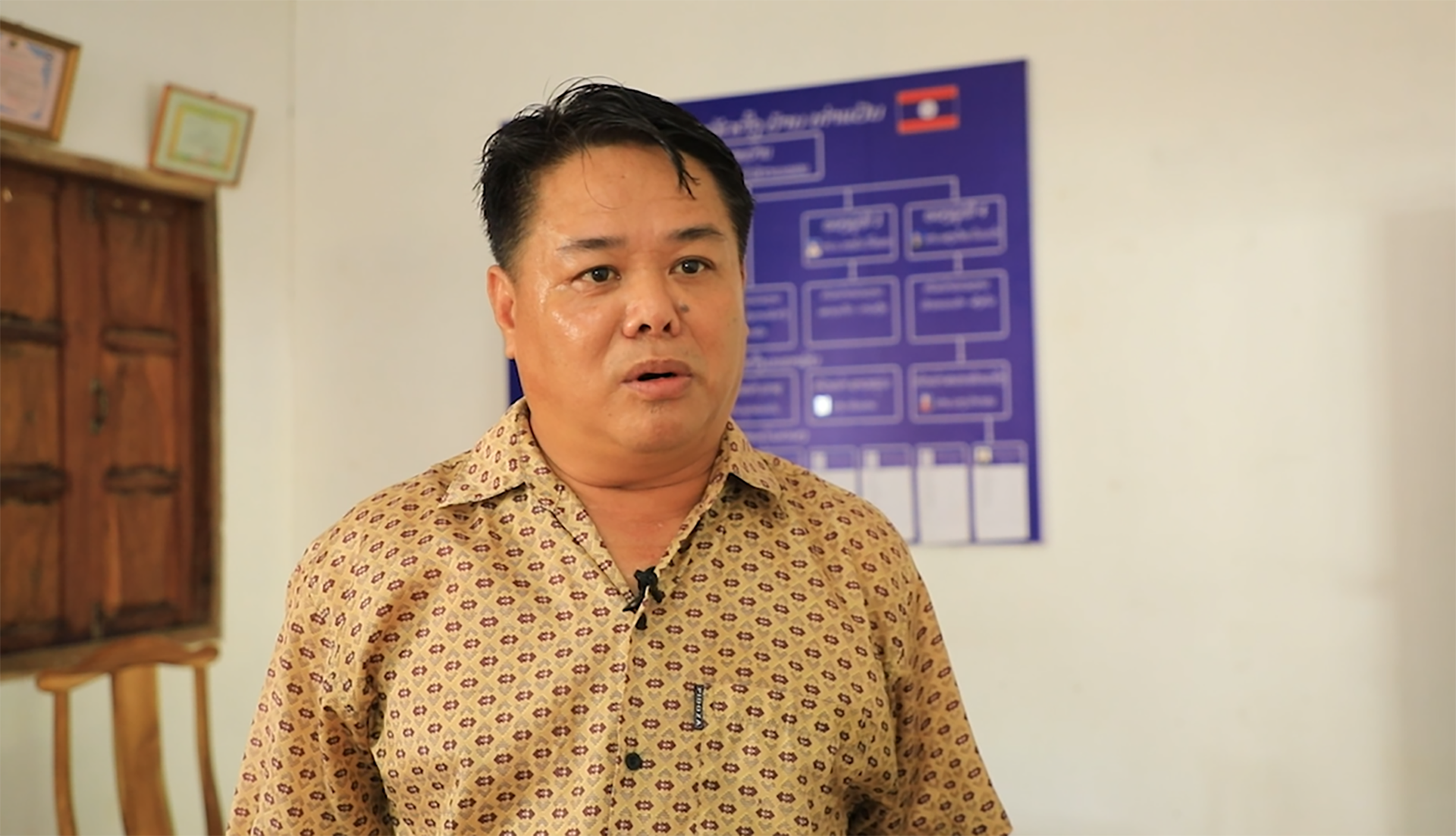 Person wearing a beige patterned shirt with a microphone attached, standing indoors near a wooden chair and door, with an organisational chart displayed on the wall in the background.