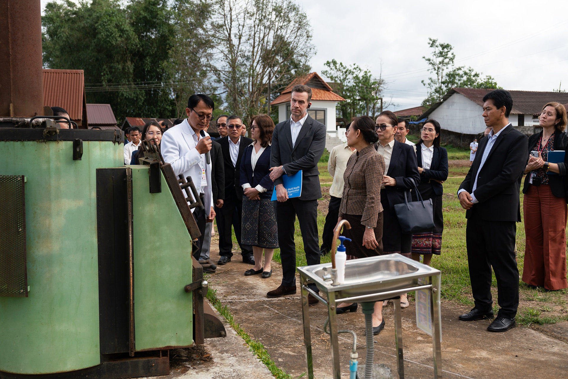 Group of people observing a demonstration of outdoor equipment near a green machine and a metal sink.