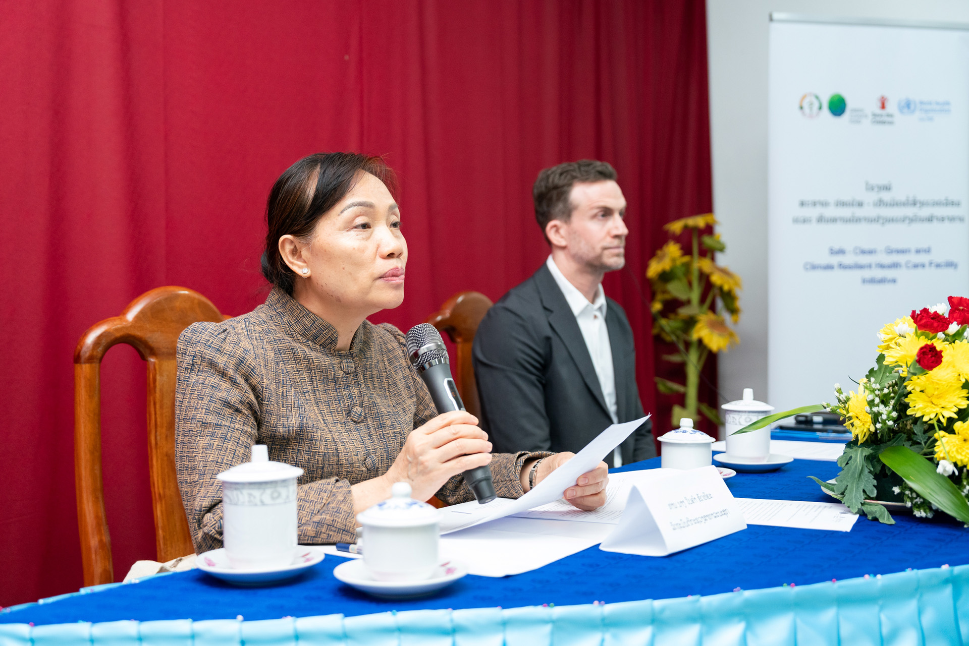 Person speaking into a microphone at a conference table with documents, tea sets, and a floral arrangement.