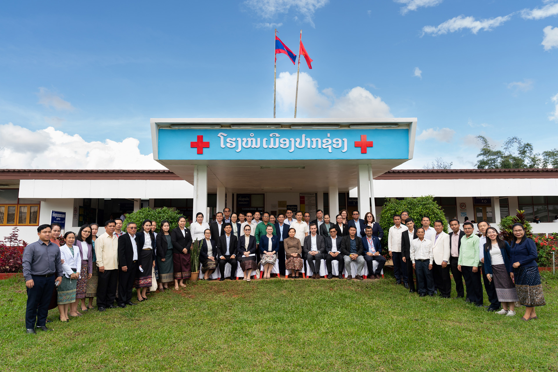 Large group posing in front of a health facility building with Lao signage and two national flags.