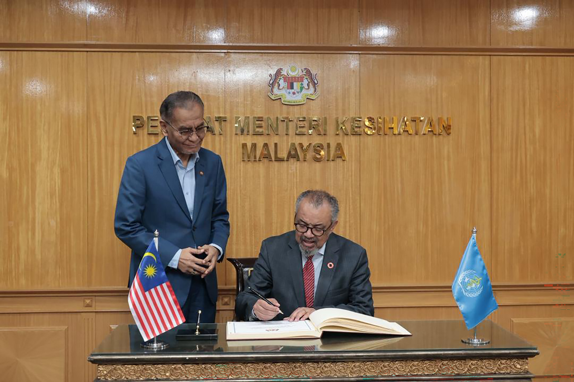 WHO Director‑General Dr Tedros Adhanom Ghebreyesus signs a guest book during an official visit to Malaysia, with a Malaysian health official standing beside him. Malaysian and WHO flags are displayed on the desk in a formal office setting.