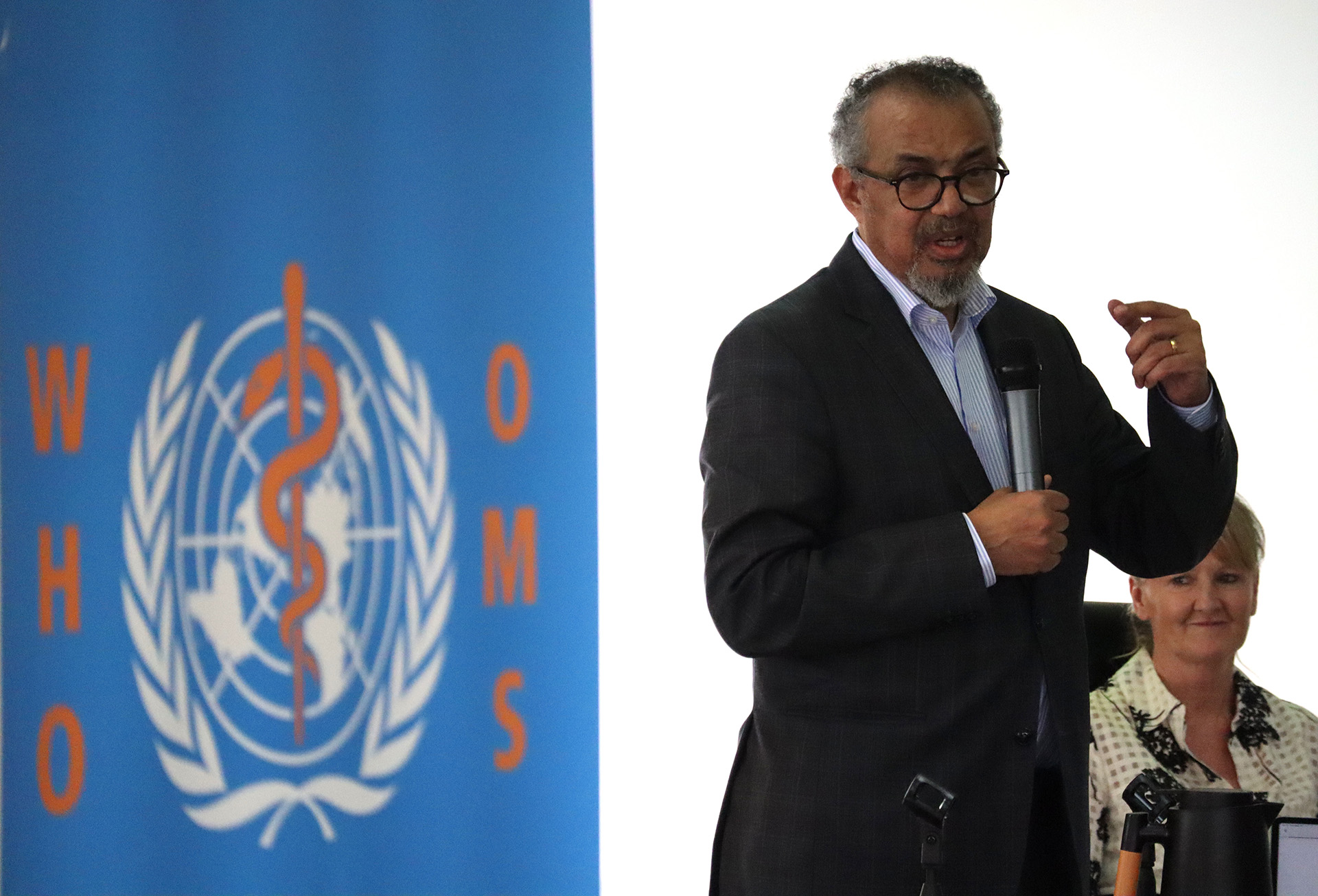 WHO Director‑General Dr Tedros Adhanom Ghebreyesus speaks into a handheld microphone during a meeting, standing beside a WHO emblem backdrop, addressing participants in a formal indoor setting.