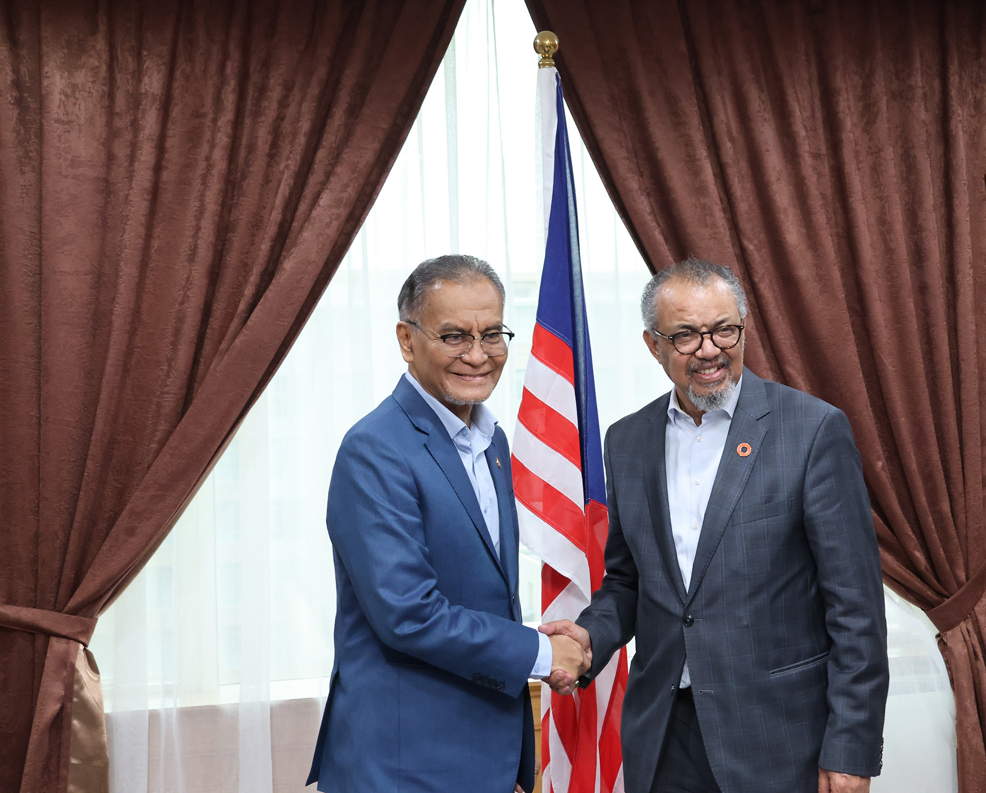 Two officials shake hands during a formal meeting indoors, standing between brown curtains with the Malaysian national flag displayed behind them, indicating a diplomatic or official engagement.
