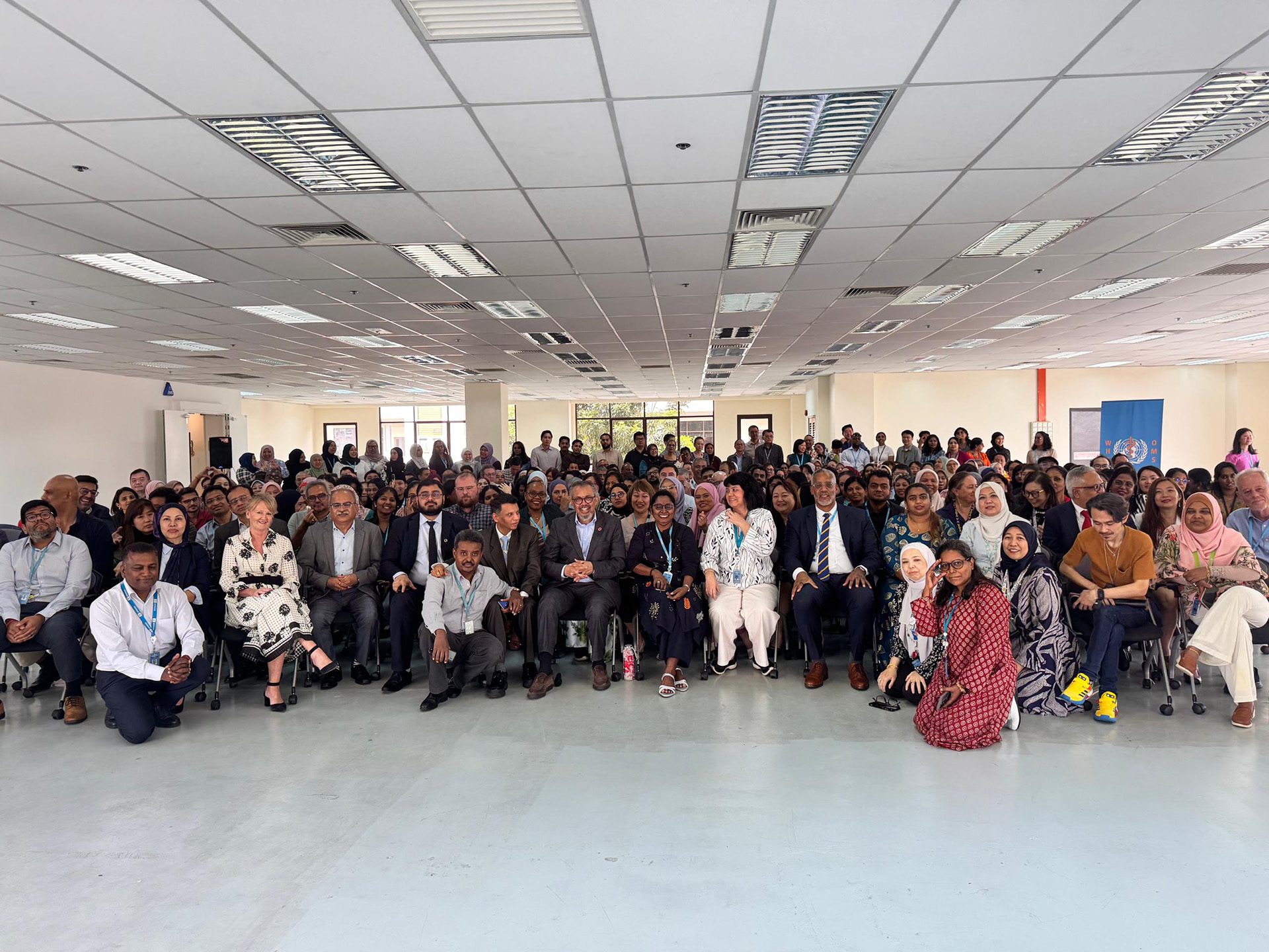 A large group of participants pose for a group photo at an indoor meeting, seated and standing together in a hall with ceiling lights, reflecting a formal gathering or collaborative event.