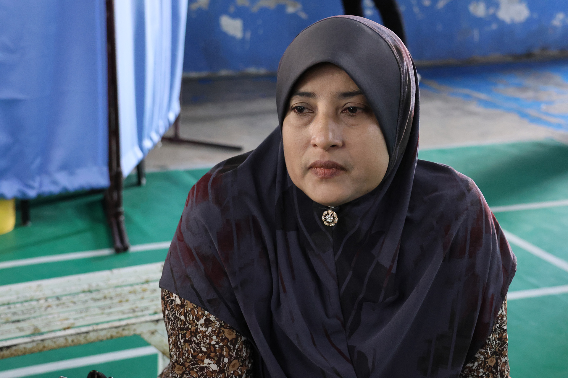 A woman wearing a dark headscarf with a decorative brooch sits indoors on a green floor,