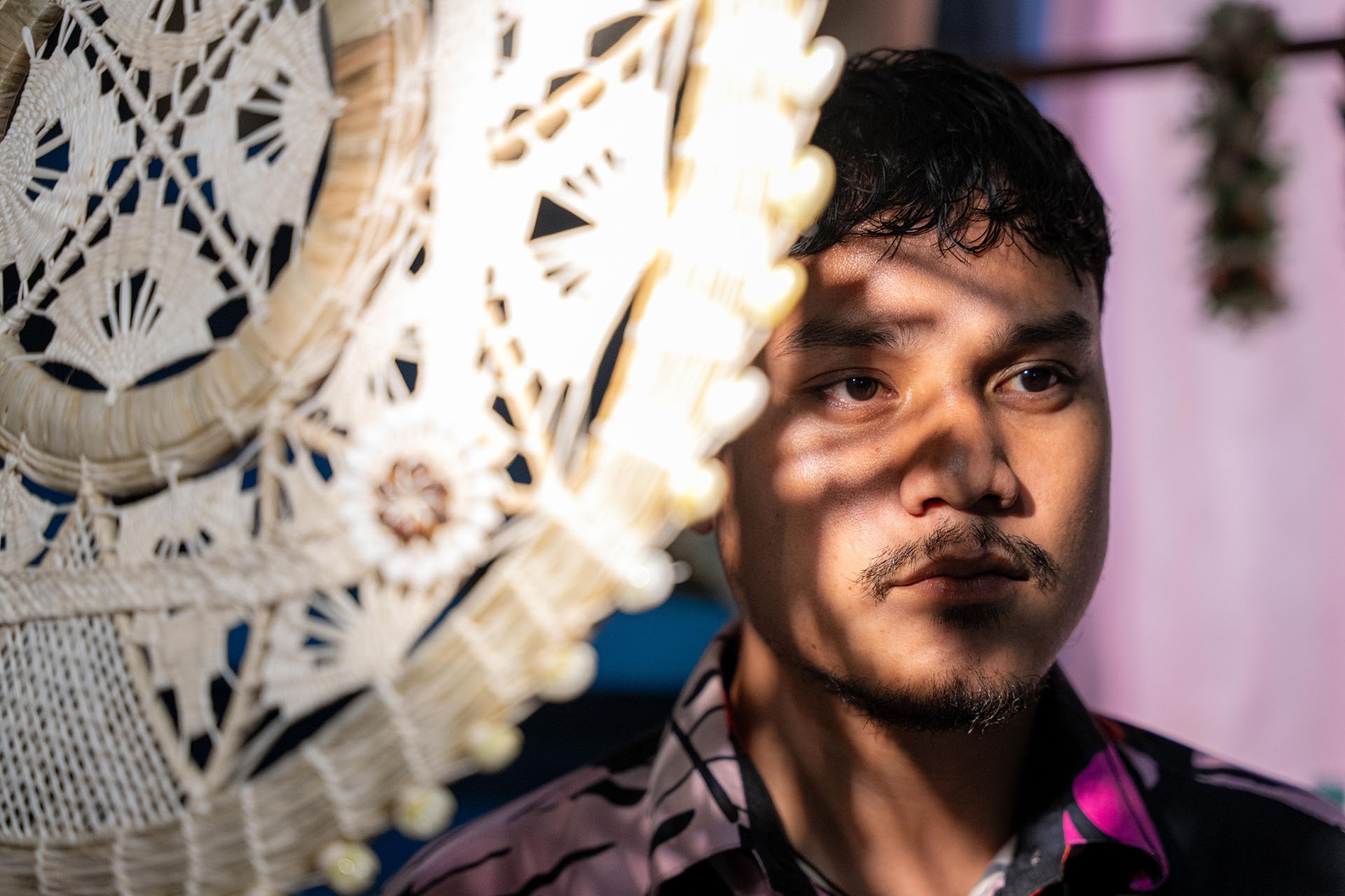 Close-up portrait of a man indoors, partially framed by lace décor, with patterned light and shadows across his face.