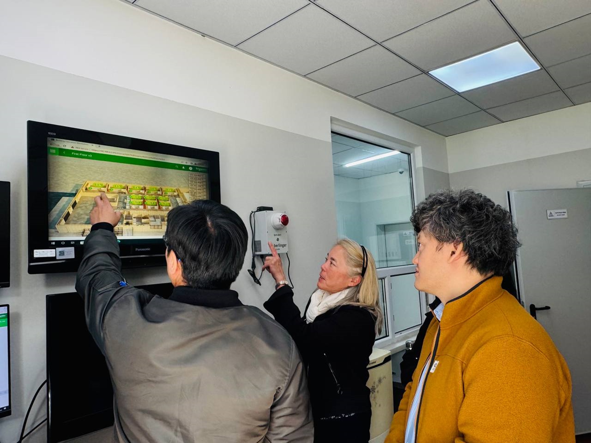 Three people point at a touchscreen showing a facility layout inside a laboratory during a site visit in Mongolia.