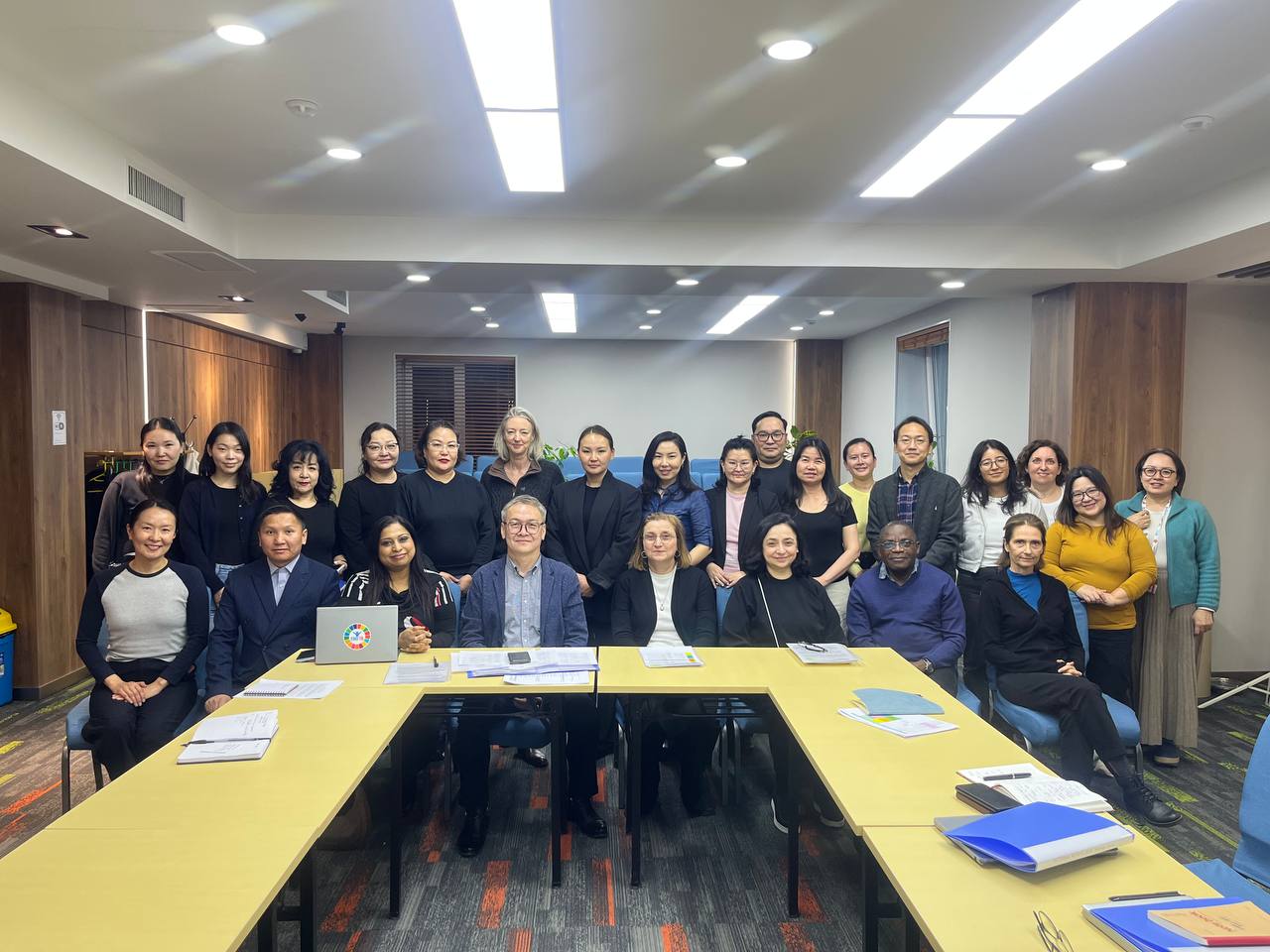 A group gathered in a meeting room, standing behind a U‑shaped conference table with laptops, papers, and folders.