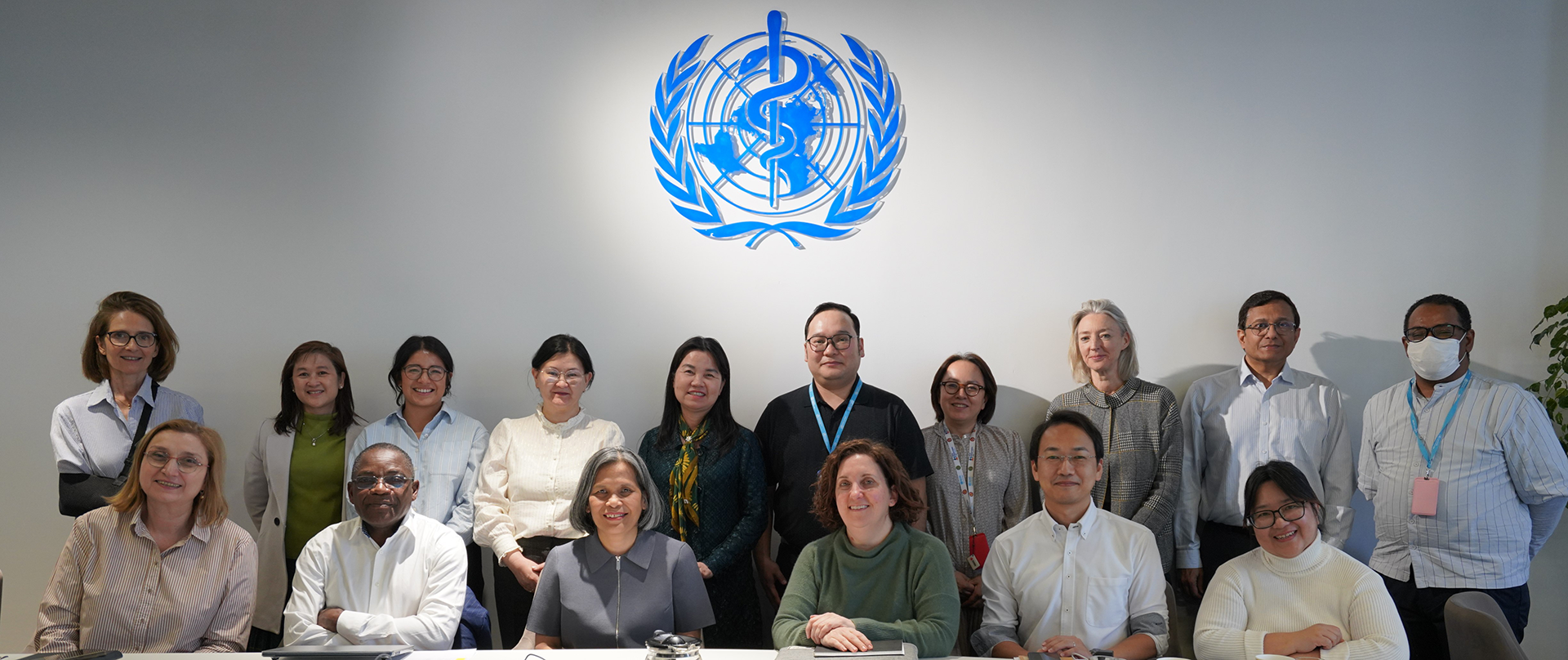 Group of international health experts seated and standing in a meeting room, posing for a group photo beneath the WHO emblem.